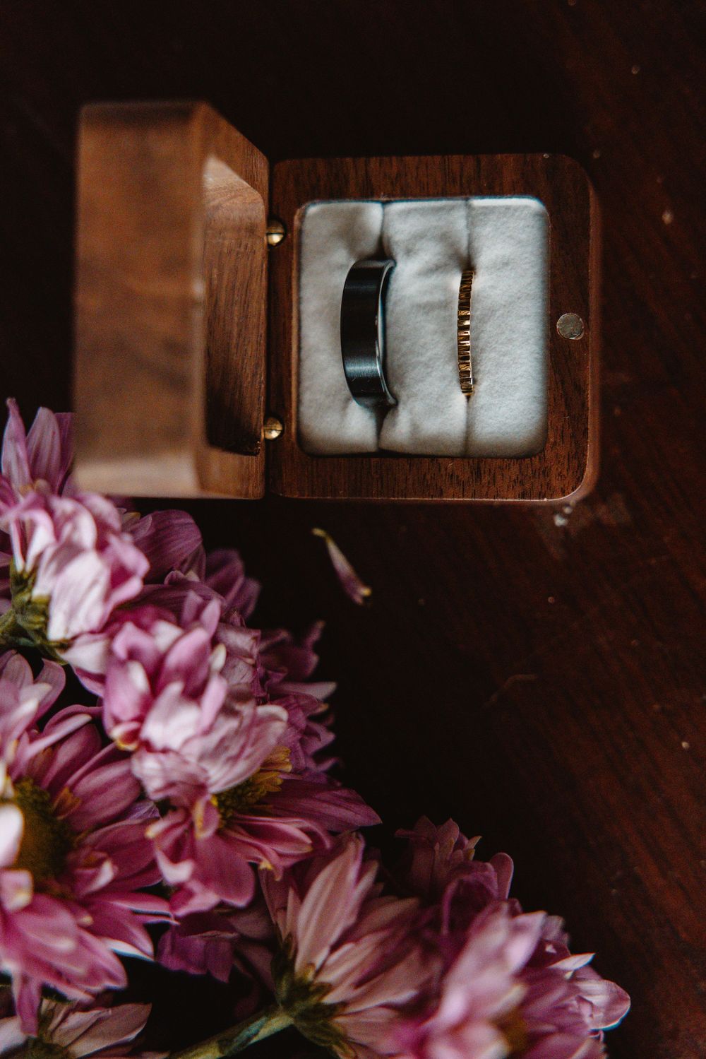 A dark tungsten wedding band displayed in an open wooden ring box surrounded by pink flowers.