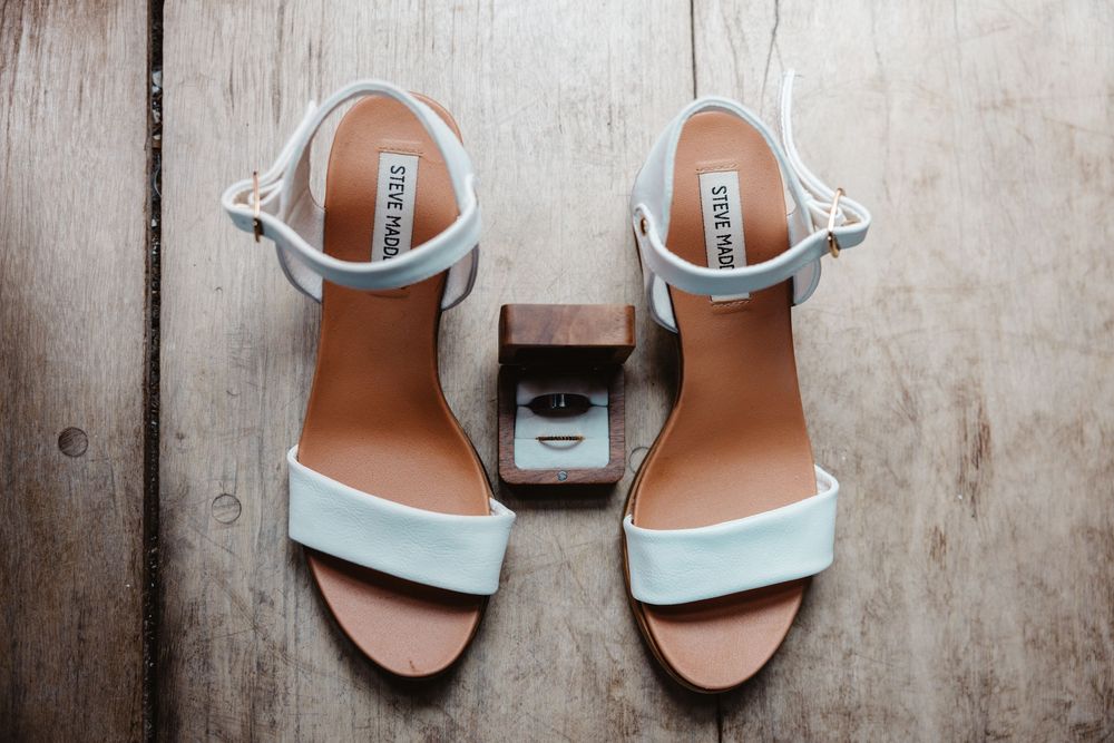 White Steve Madden heeled sandals displayed on wood with a small wooden ring box between them.