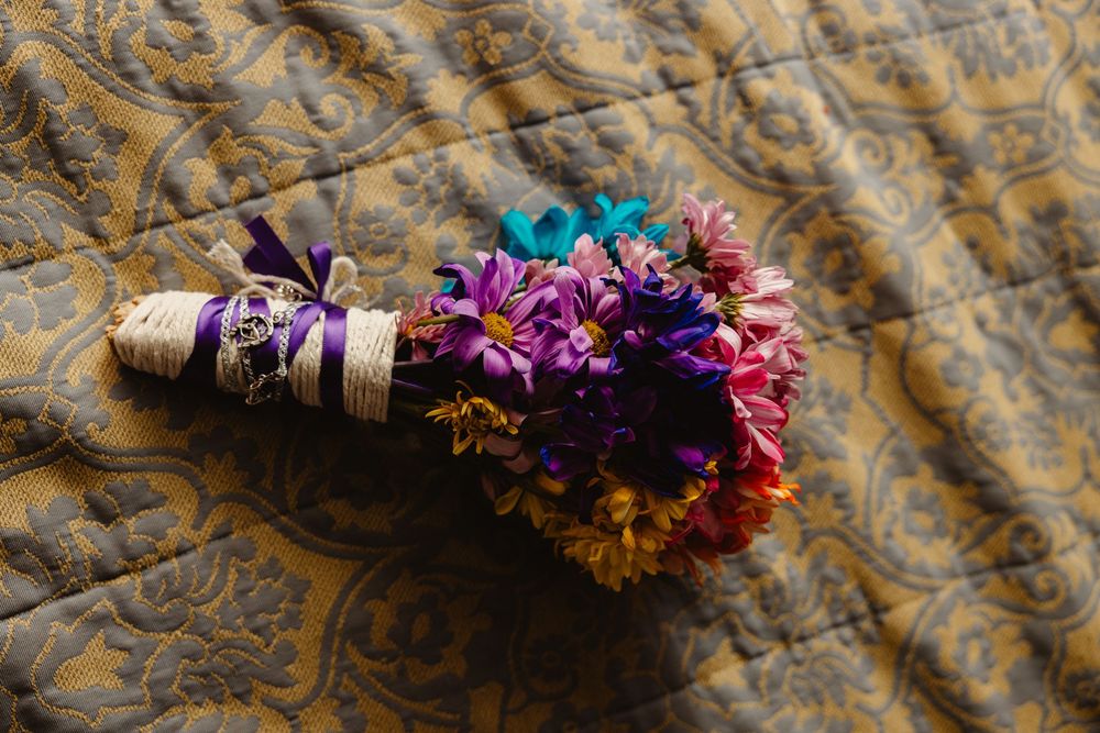 A colorful bridal bouquet with purple, blue, and orange flowers wrapped in lace ribbon on a quilt.
