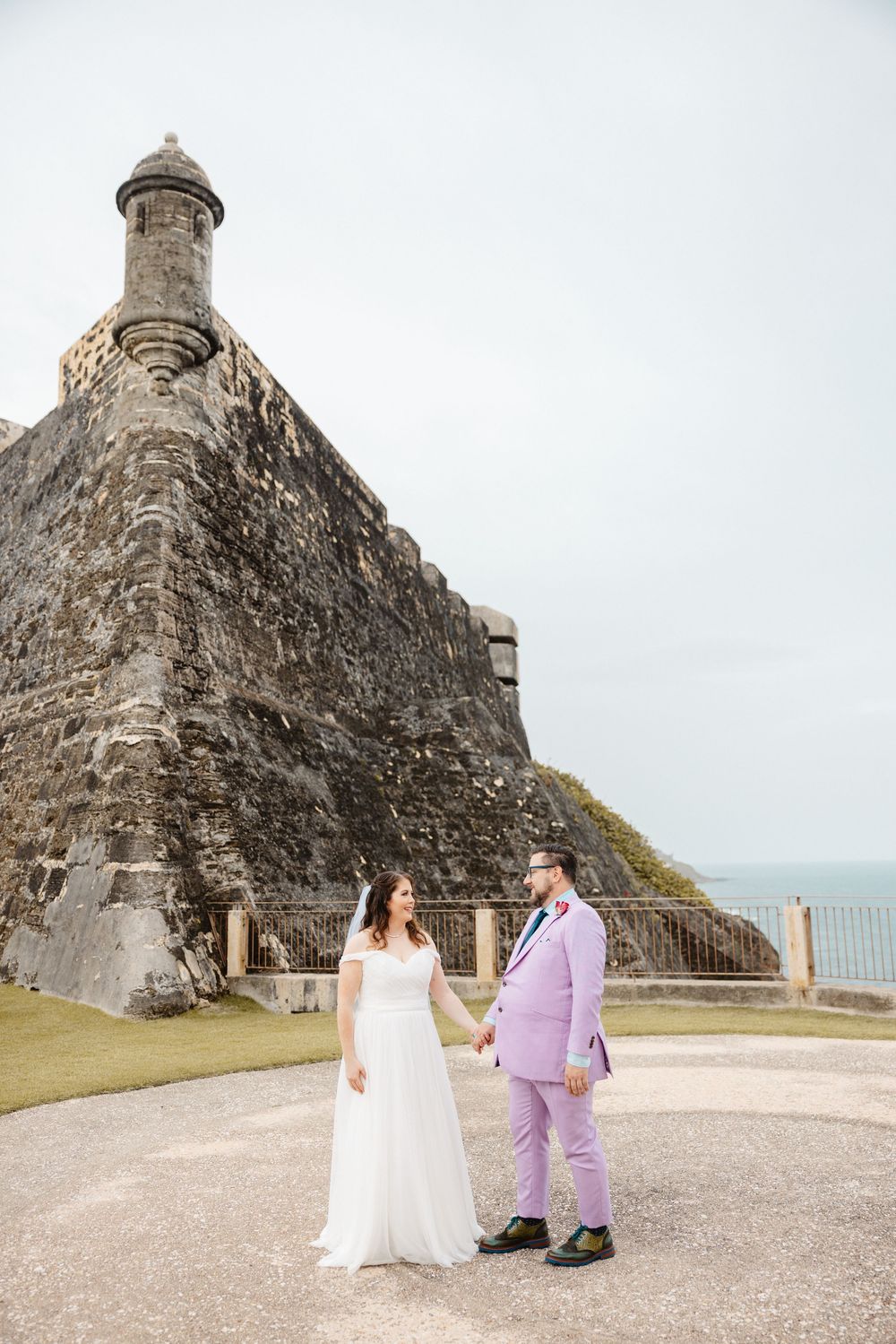 A bride and groom hold hands in front of a historic stone fortress with ocean views.