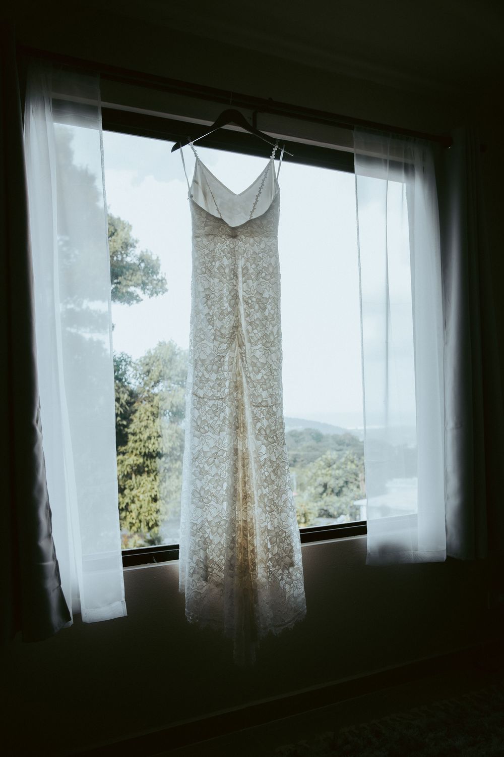 Elegant lace wedding dress hanging in window with sheer curtains and lush green view.