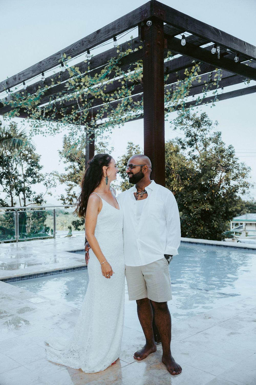 Interracial wedding couple standing by rooftop pool under pergola with string lights overhead.