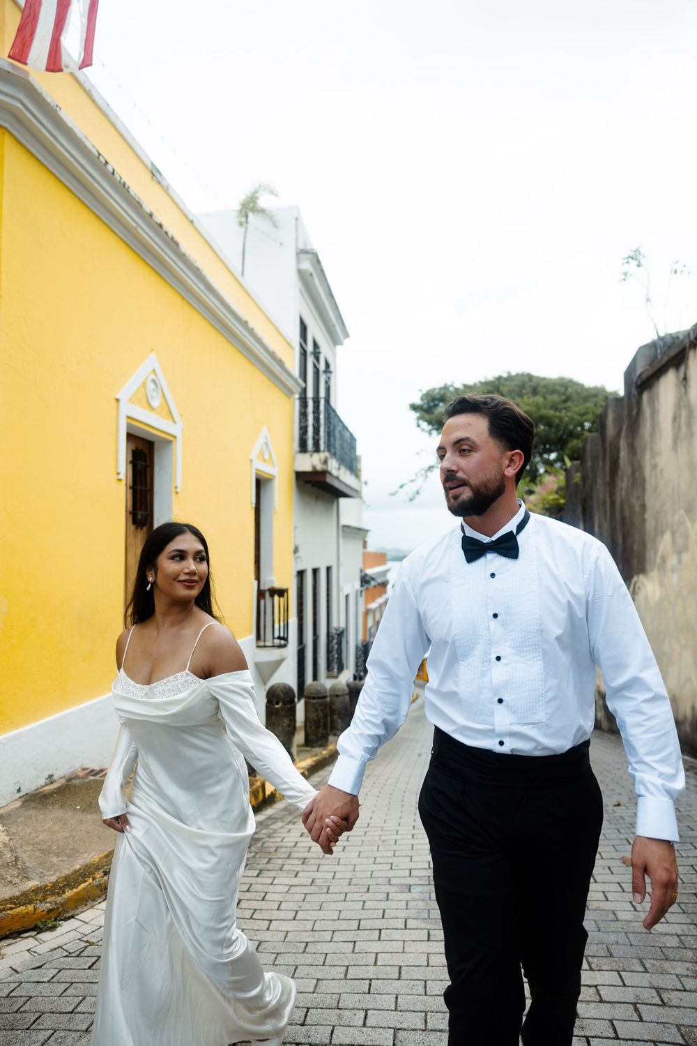 Bride and groom walking hand in hand on cobblestone street in Old San Juan, Puerto Rico, with colorful buildings.