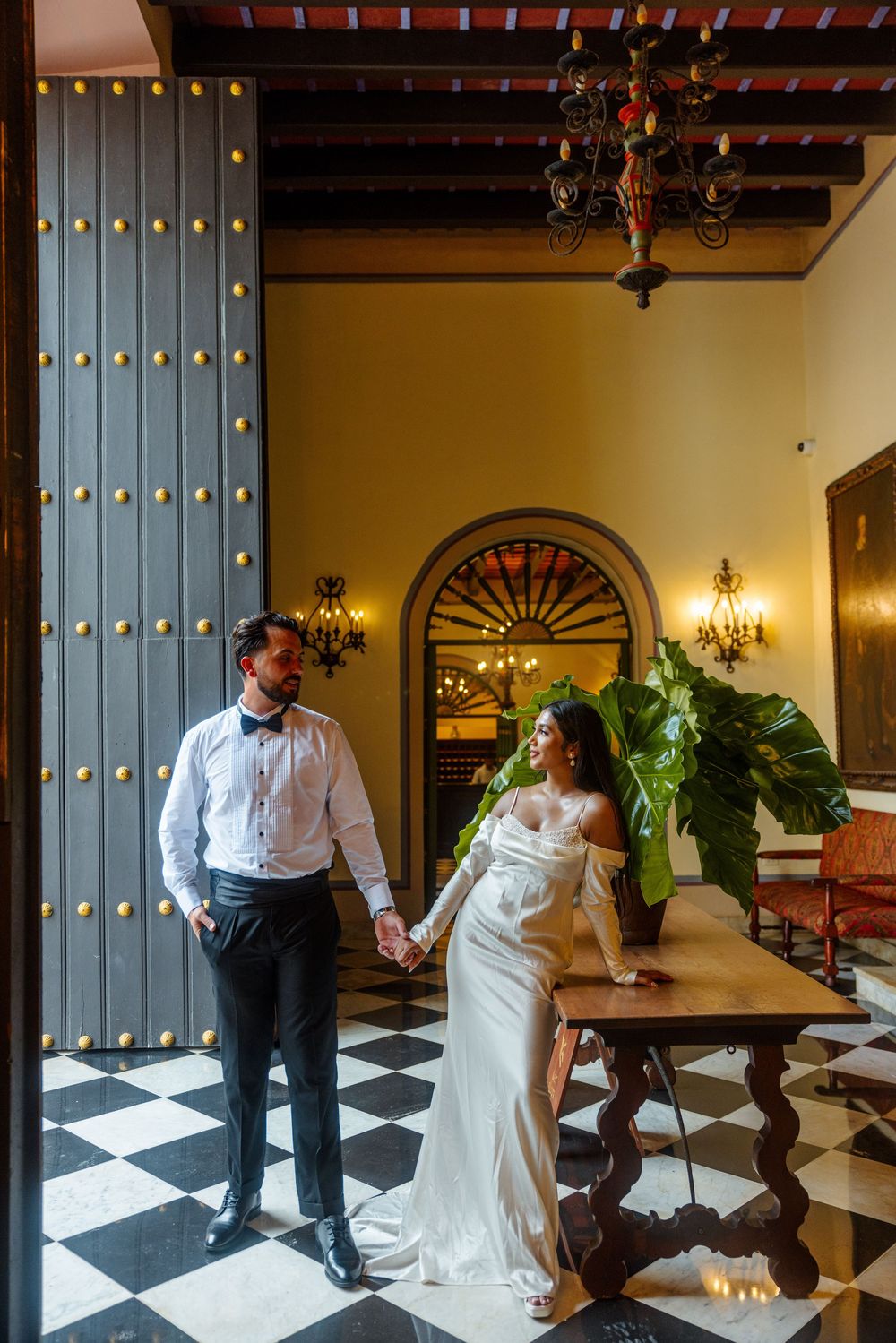 Bride and groom holding hands in elegant colonial venue with checkered floors and ornate wooden doors.