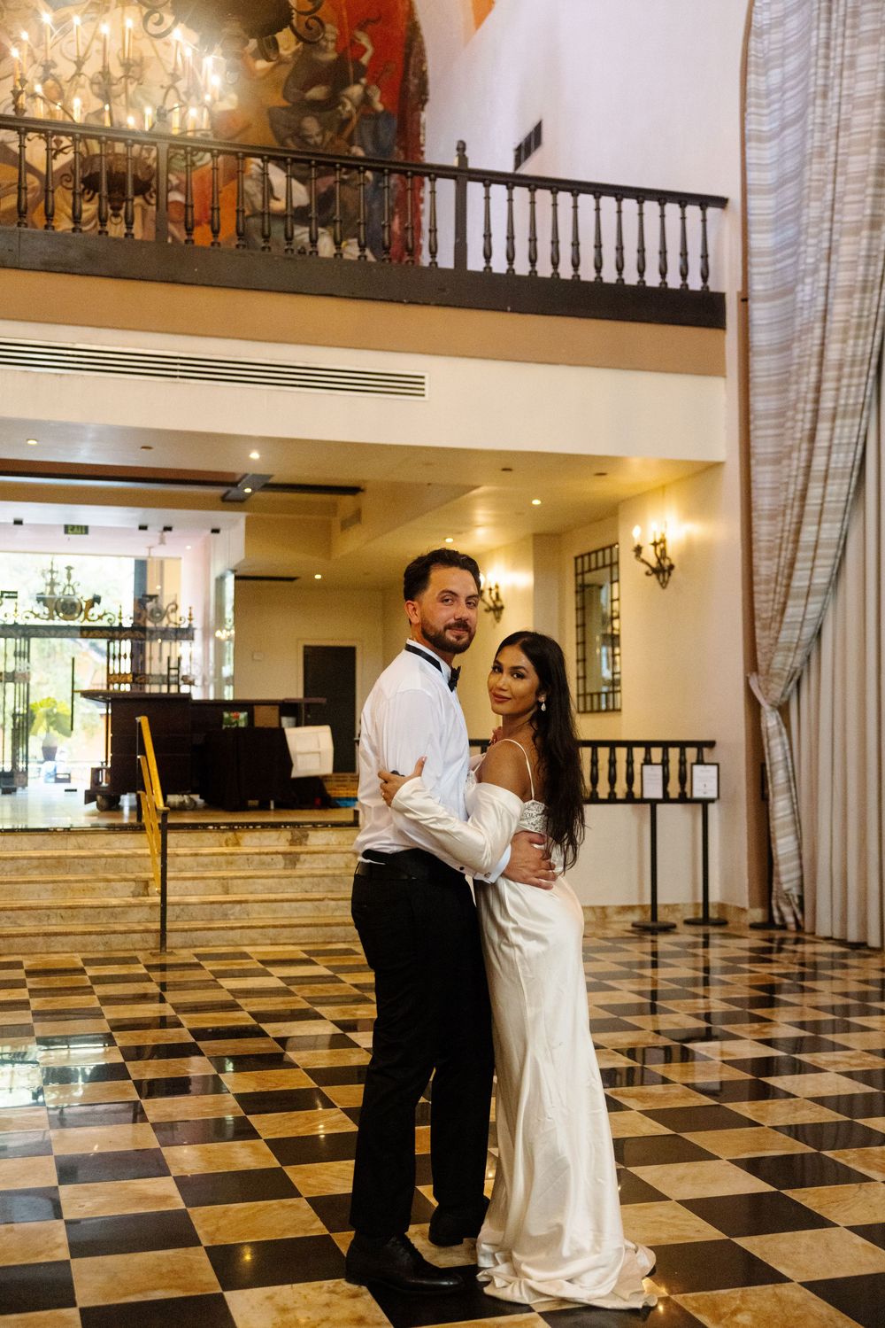 Wedding couple embracing in luxurious hotel lobby with checkered marble floors and grand balcony.