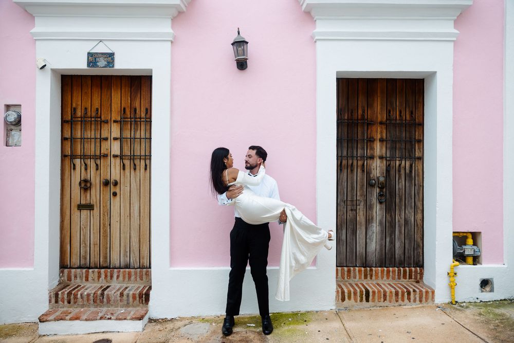 Groom lifting bride in white dress against vibrant pink colonial building with wooden doors.