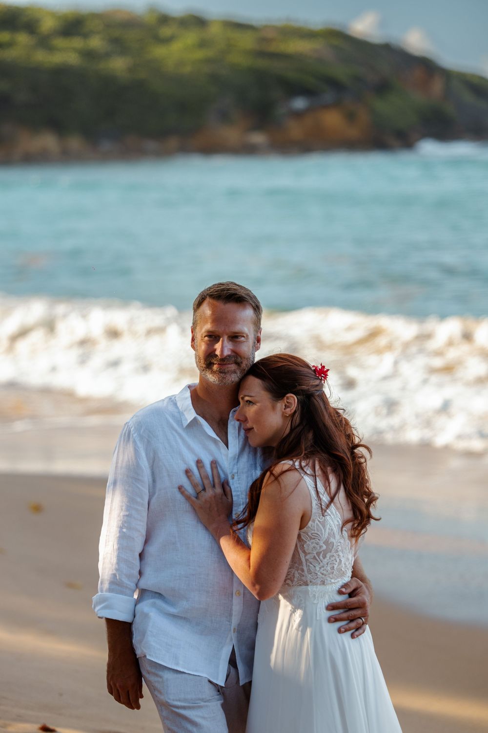 Beach wedding couple embracing on sandy shore with turquoise ocean waves in the background.