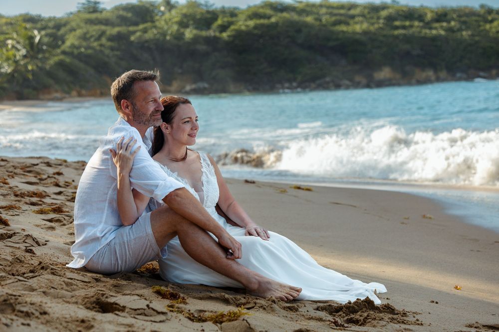 Newlyweds sitting together on sandy beach gazing at ocean waves during golden hour.