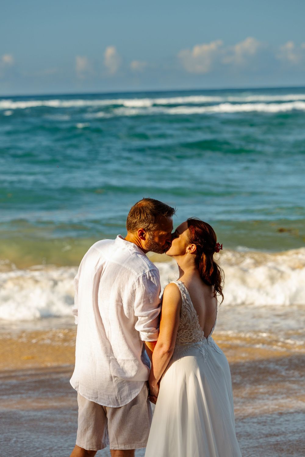 Bride and groom sharing a romantic kiss on the beach with crashing waves behind them.