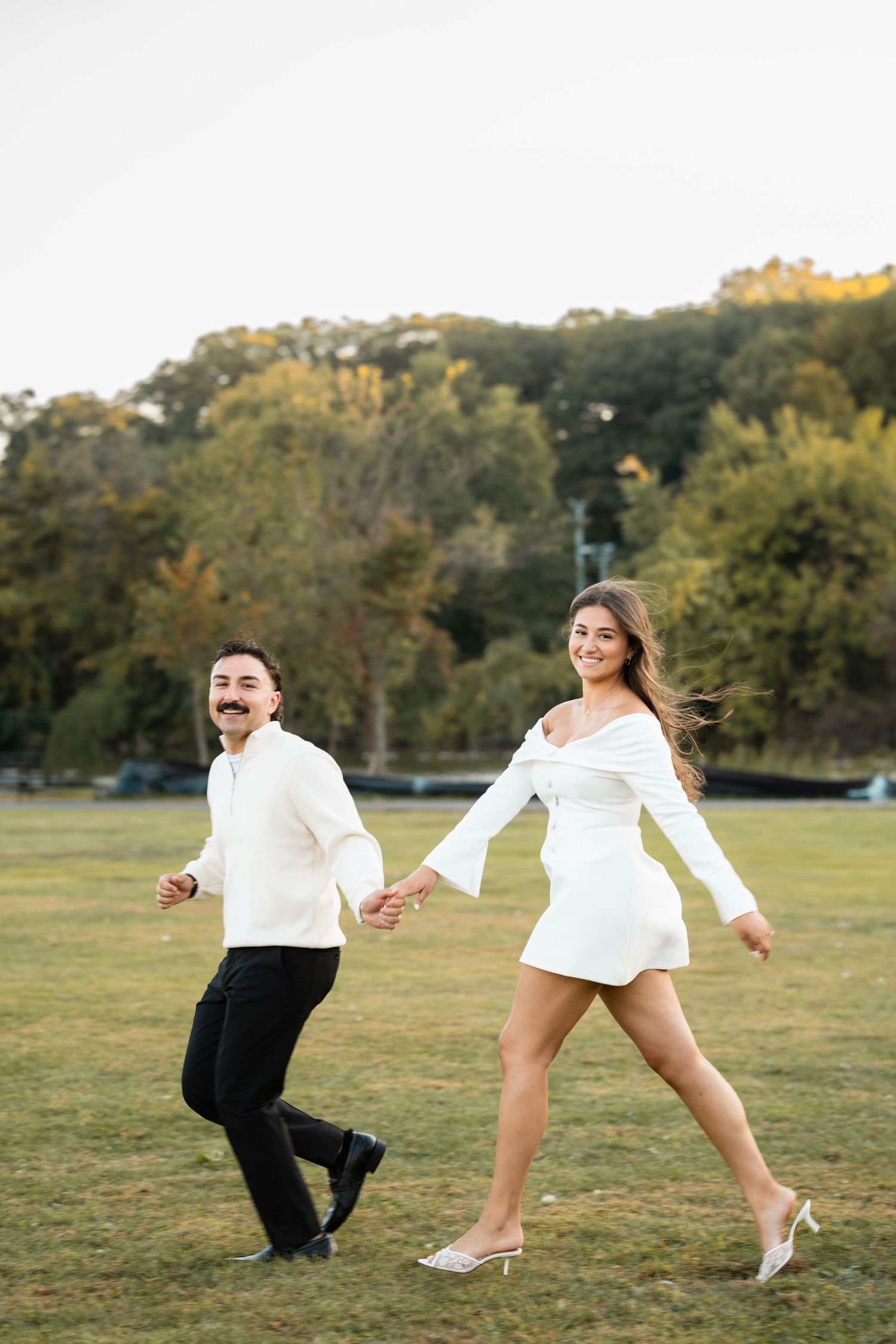 Playful couple in formal wear jumping and dancing on grassy field at sunset.