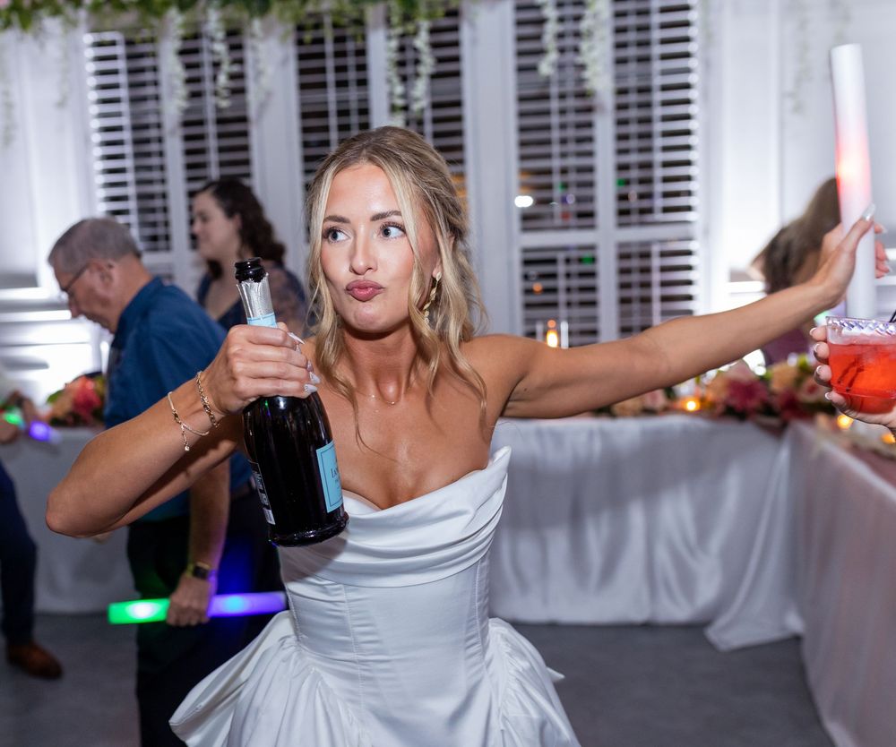 Bride in white strapless gown dancing at wedding reception holding champagne bottle with arm outstretched.