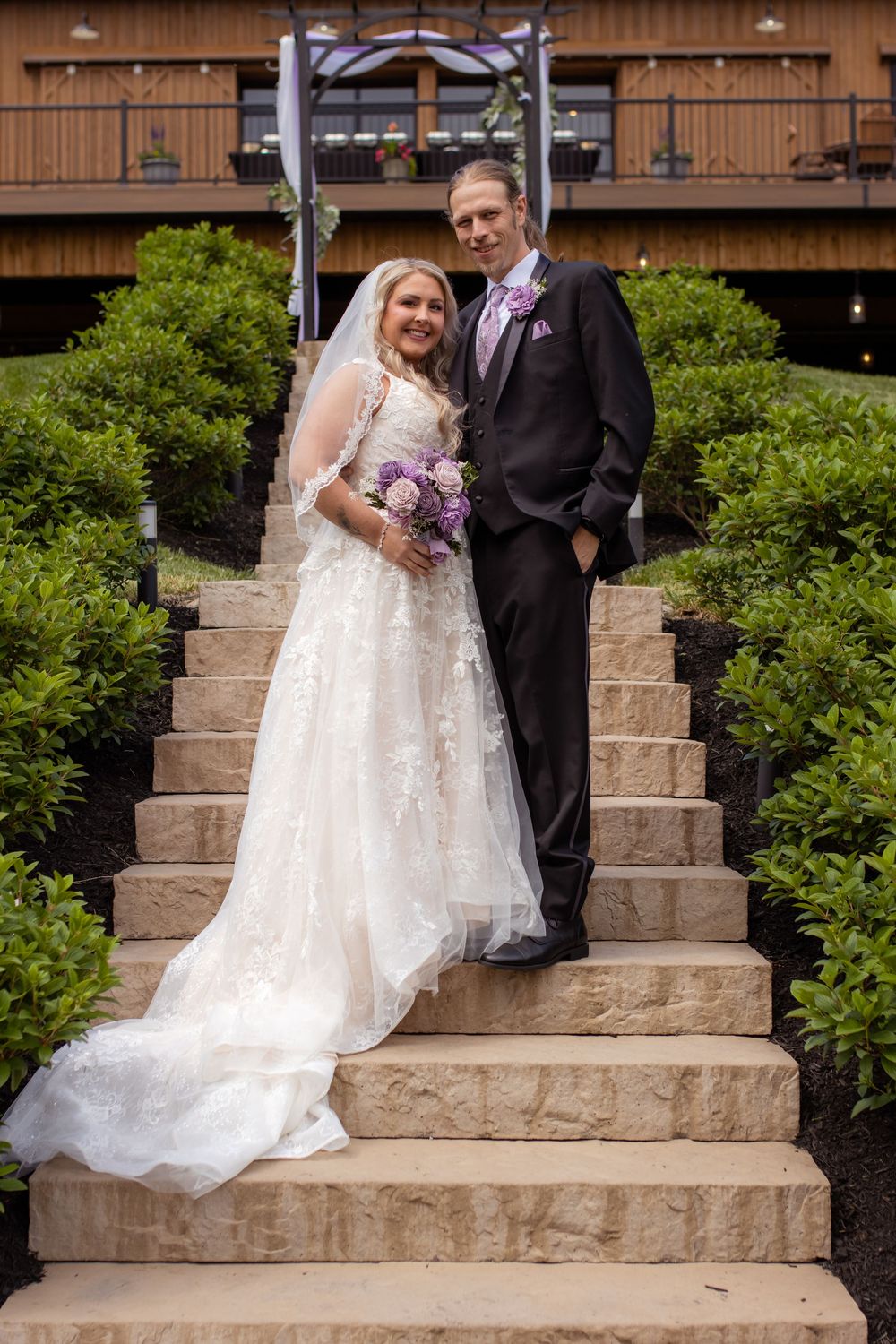 A couple stands together on stone steps in front of a rustic wooden lodge on their wedding day, surrounded by green shrubs.