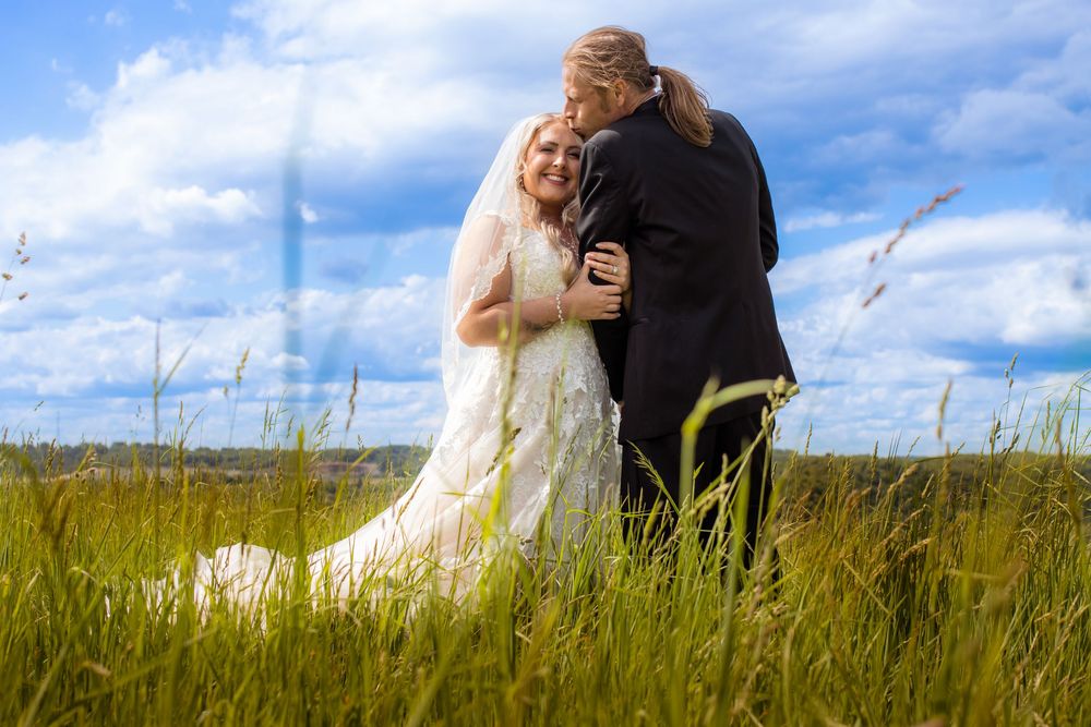 A couple embraces in a serene meadow with tall grass under a blue cloudy sky during a wedding photoshoot.