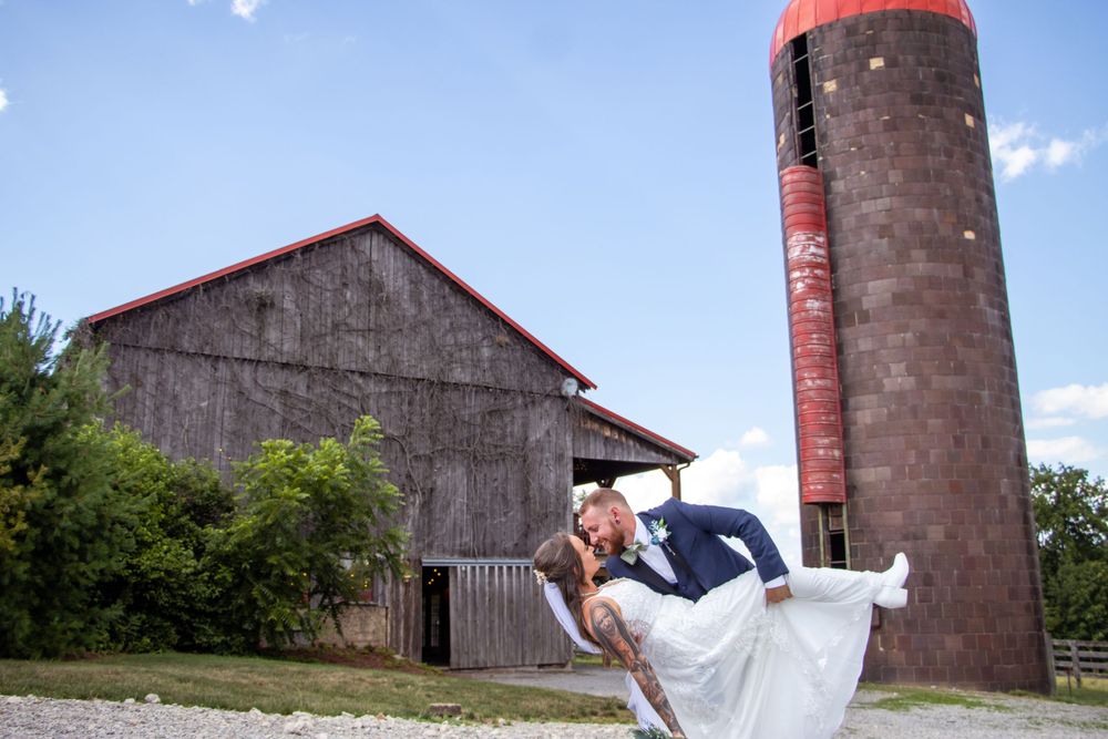 Couple in wedding attire share romantic moment in front of rustic barn and red silo on sunny day.