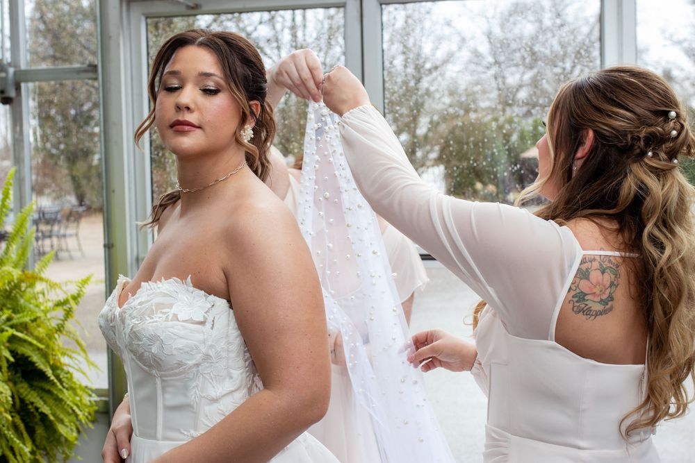 A bride prepares for her wedding day as someone helps adjust the finishing touches of her white bridal gown.