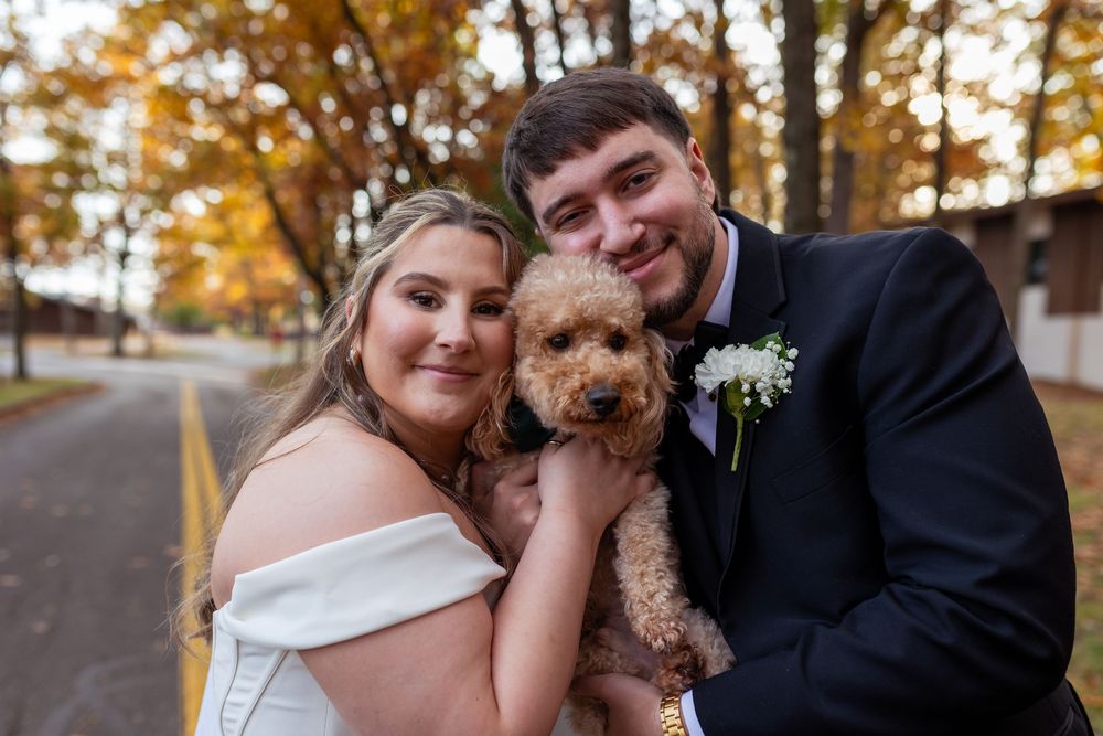 Bride and groom holding a fluffy golden doodle puppy outdoors in autumn, smiling together in wedding attire.