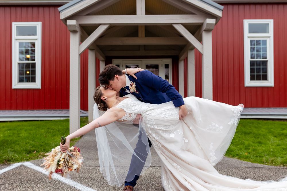 Groom dips bride in romantic kiss outside red barn wedding venue, bride holding colorful bouquet in flowing white gown.