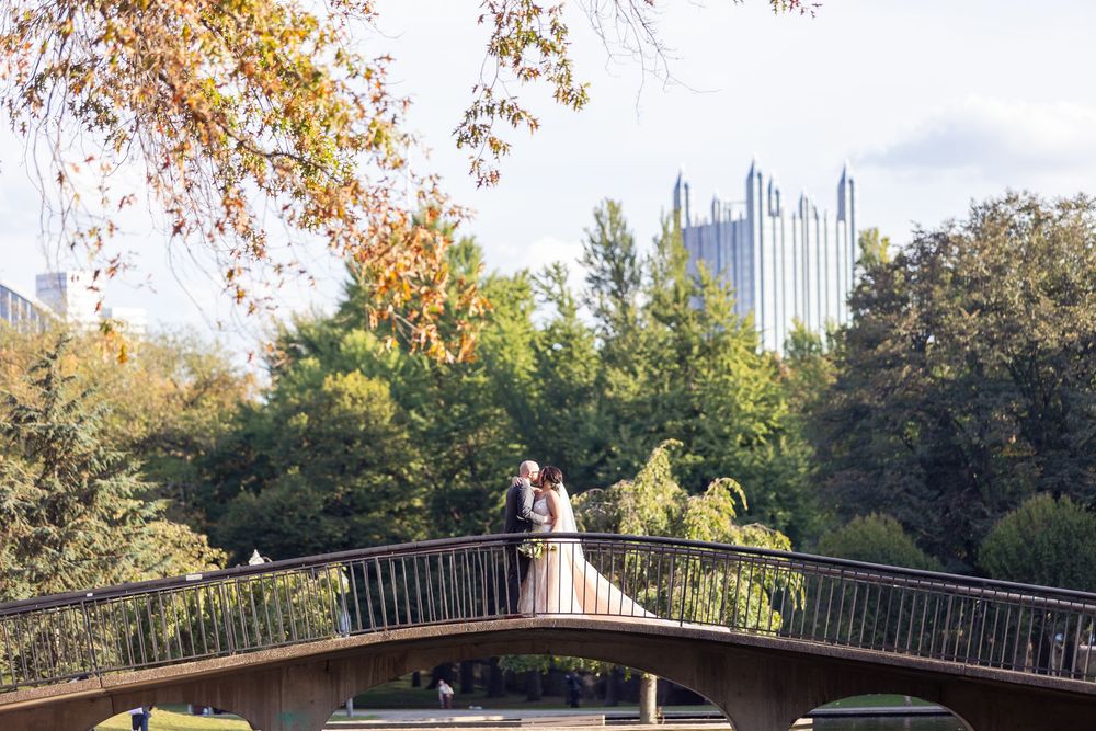 Bride and groom kissing on a wooden bridge in Pittsburgh with city skyline and autumn trees in background.