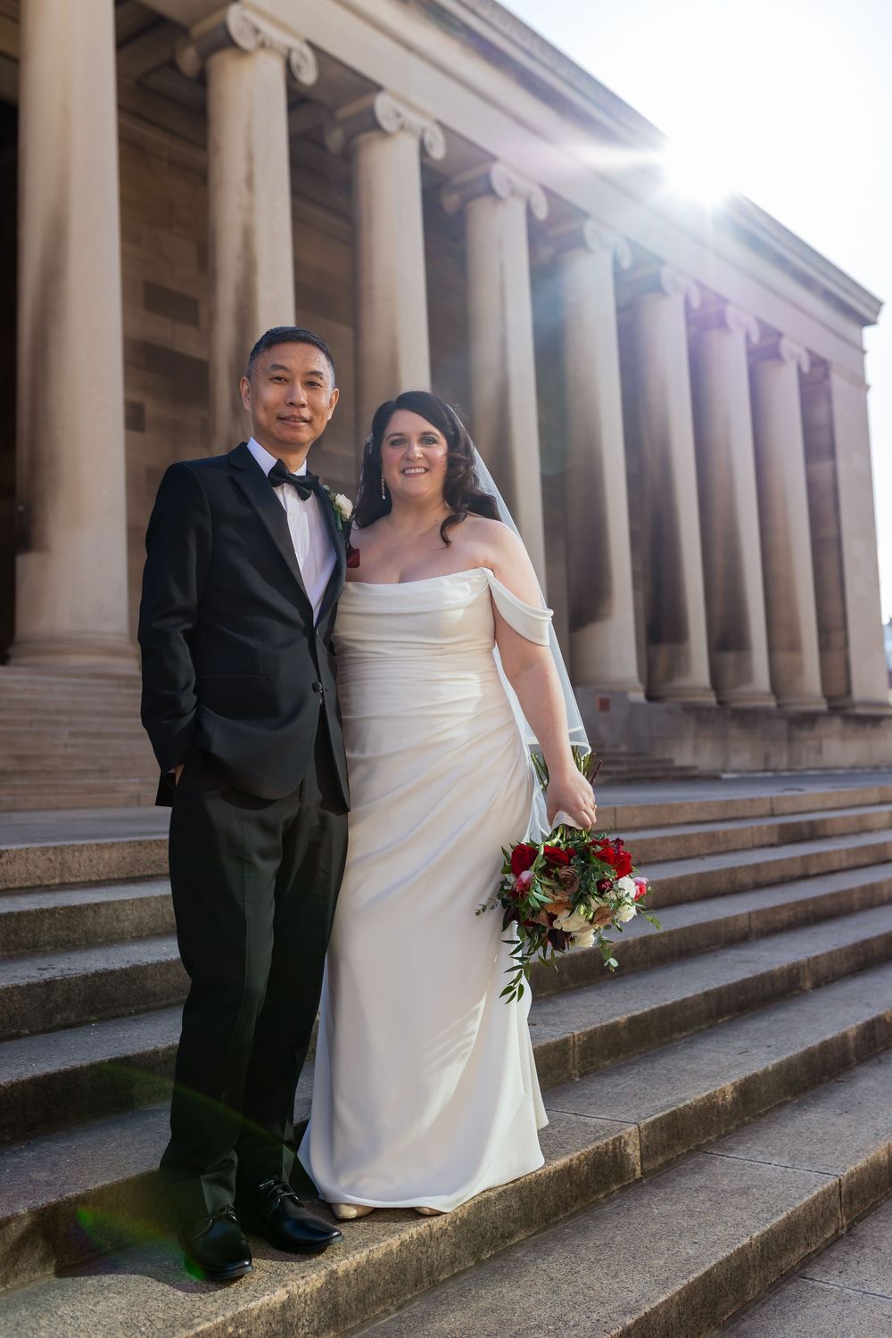 Bride and groom posing on stone steps of a classical building with columns, bride holding red and white bouquet.