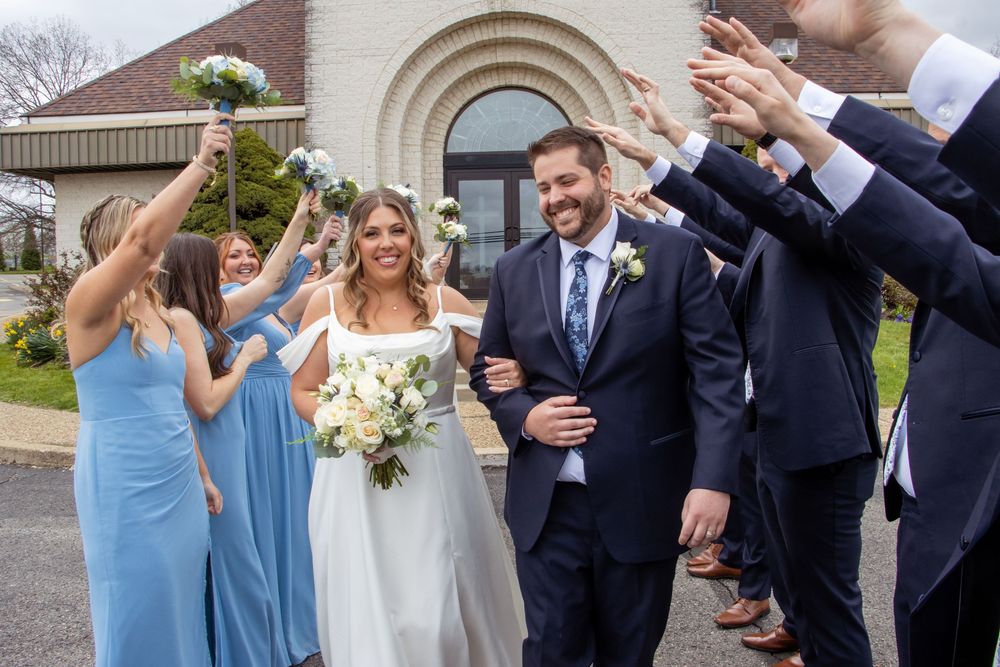 Wedding party celebrating with outstretched arms as couple exits church ceremony in blue and white attire.