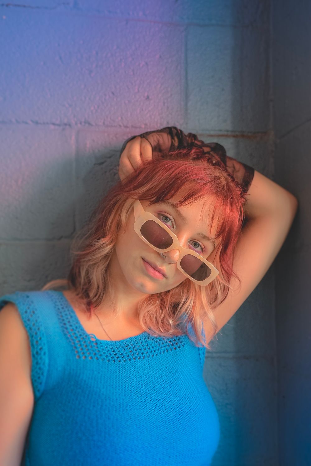 Portrait in blue top with sunglasses and pink hair against wall with colored lighting.