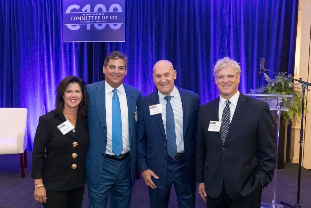 Business professionals pose for group photo at conference with C100 banner in background.