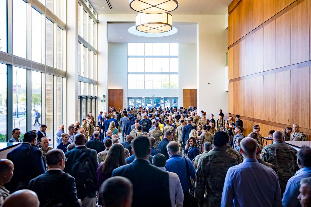 Large crowd of formally dressed attendees gather in modern venue with floor-to-ceiling windows.