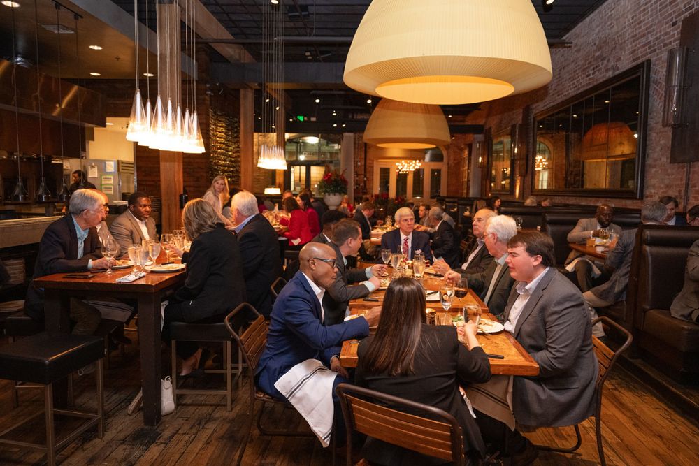 Busy restaurant interior with groups of diners seated at wooden tables under warm lighting.