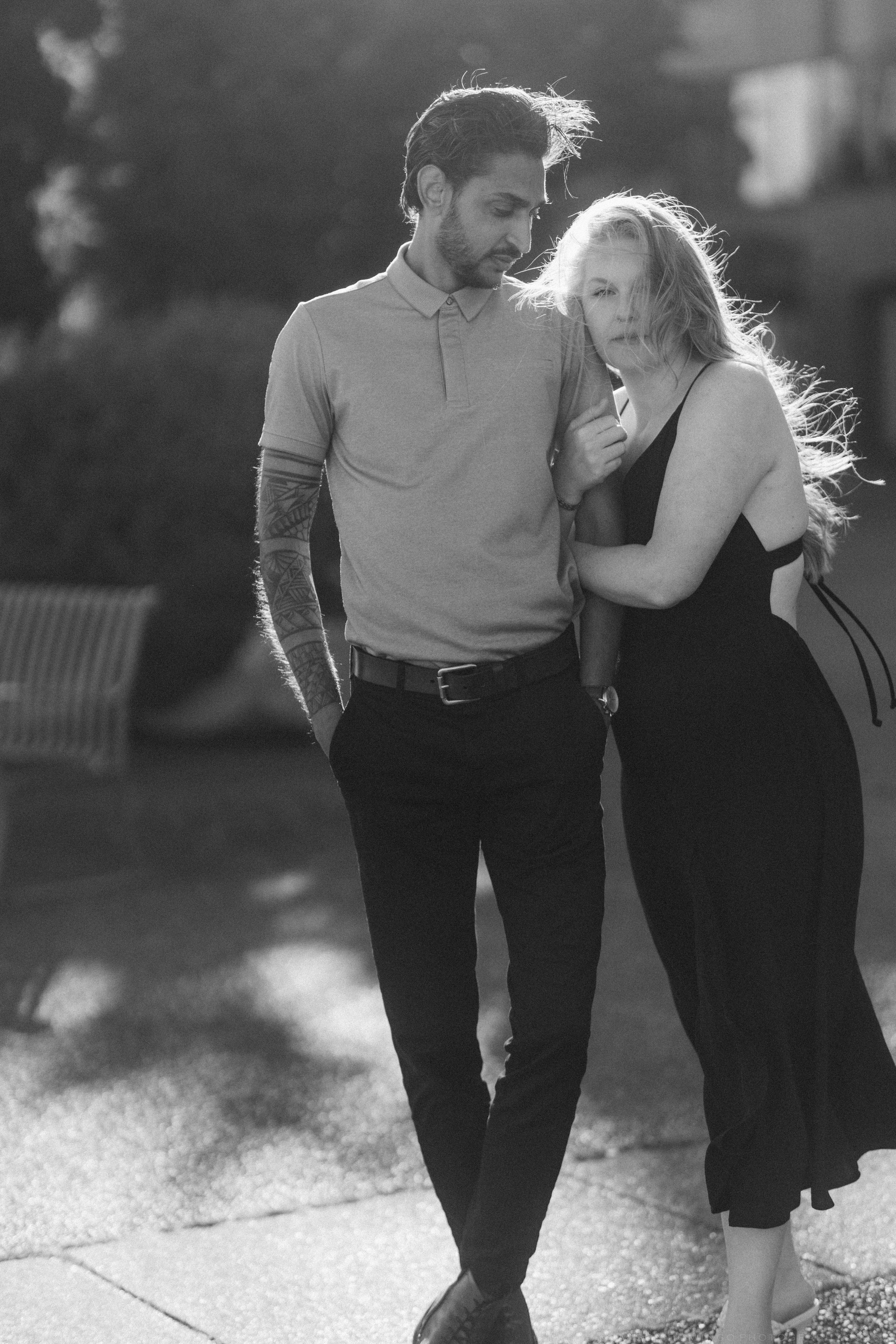 Black and white photo of a couple walking together outdoors, woman leaning on mans arm, wind blowing her hair.