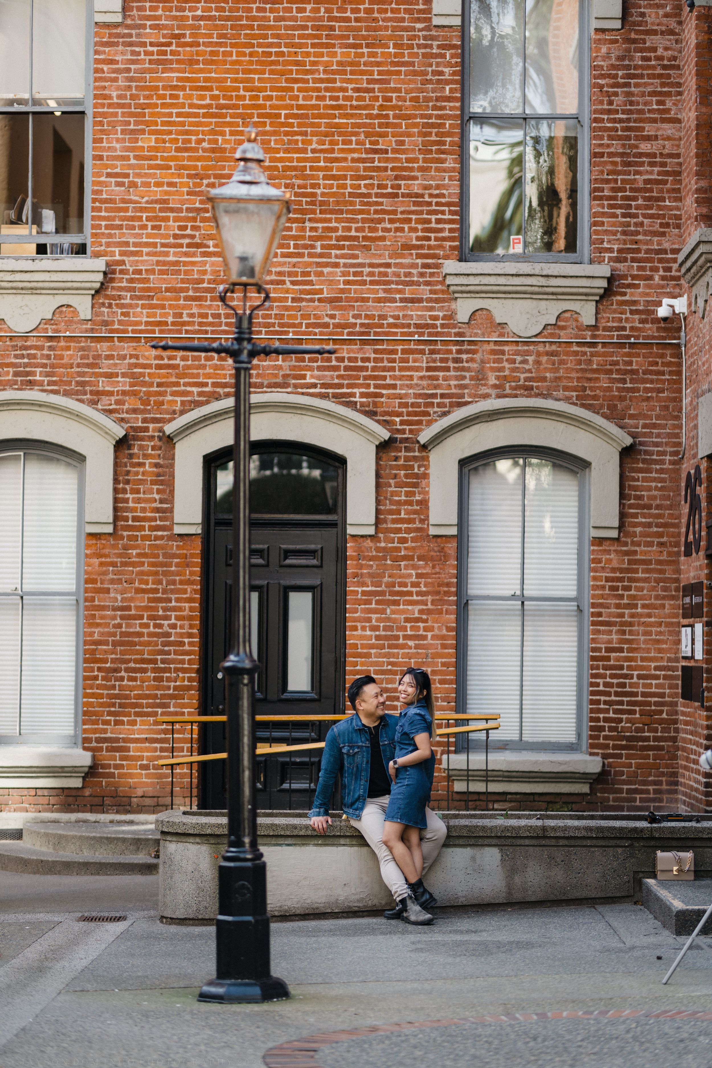 Couple embracing by a vintage street lamp in front of a red brick building with arched windows.