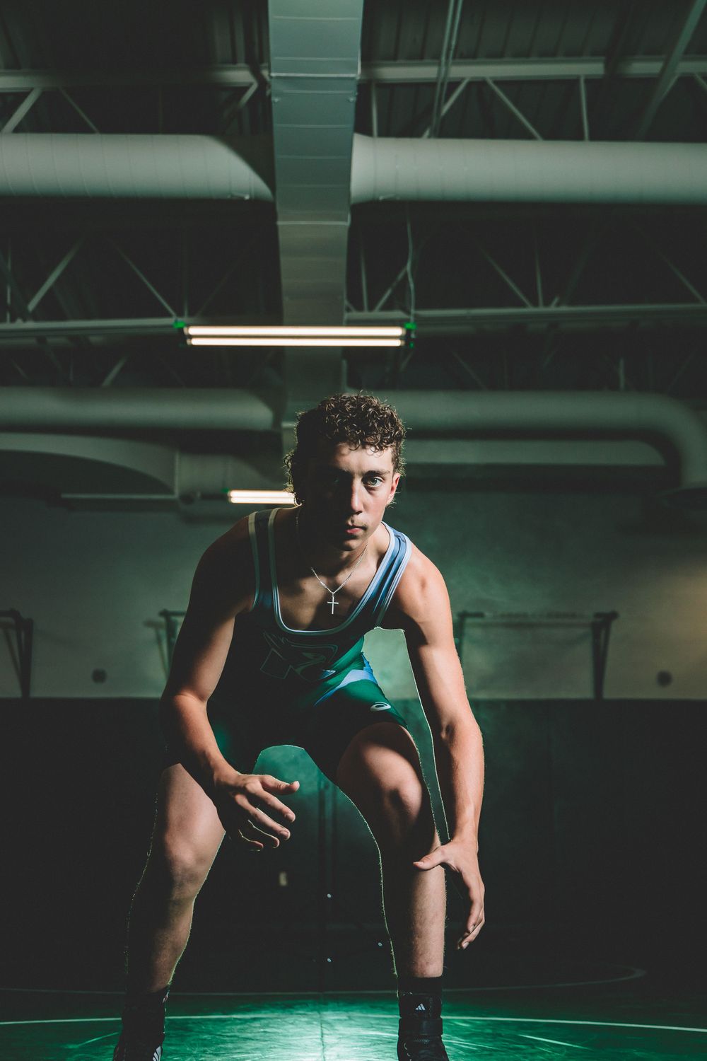 Focused Riverside High School wrestler in a green singlet assumes a ready stance on a dimly lit mat, prepared for competition