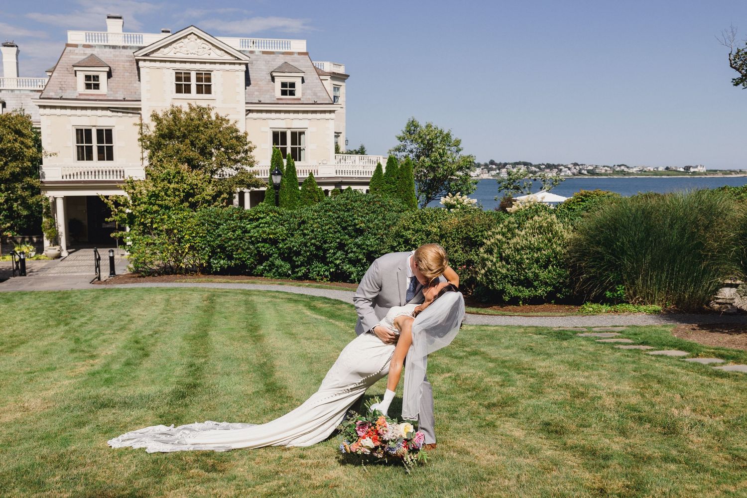 A stately white mansion with manicured lawn overlooks a waterfront view on a sunny summer day.