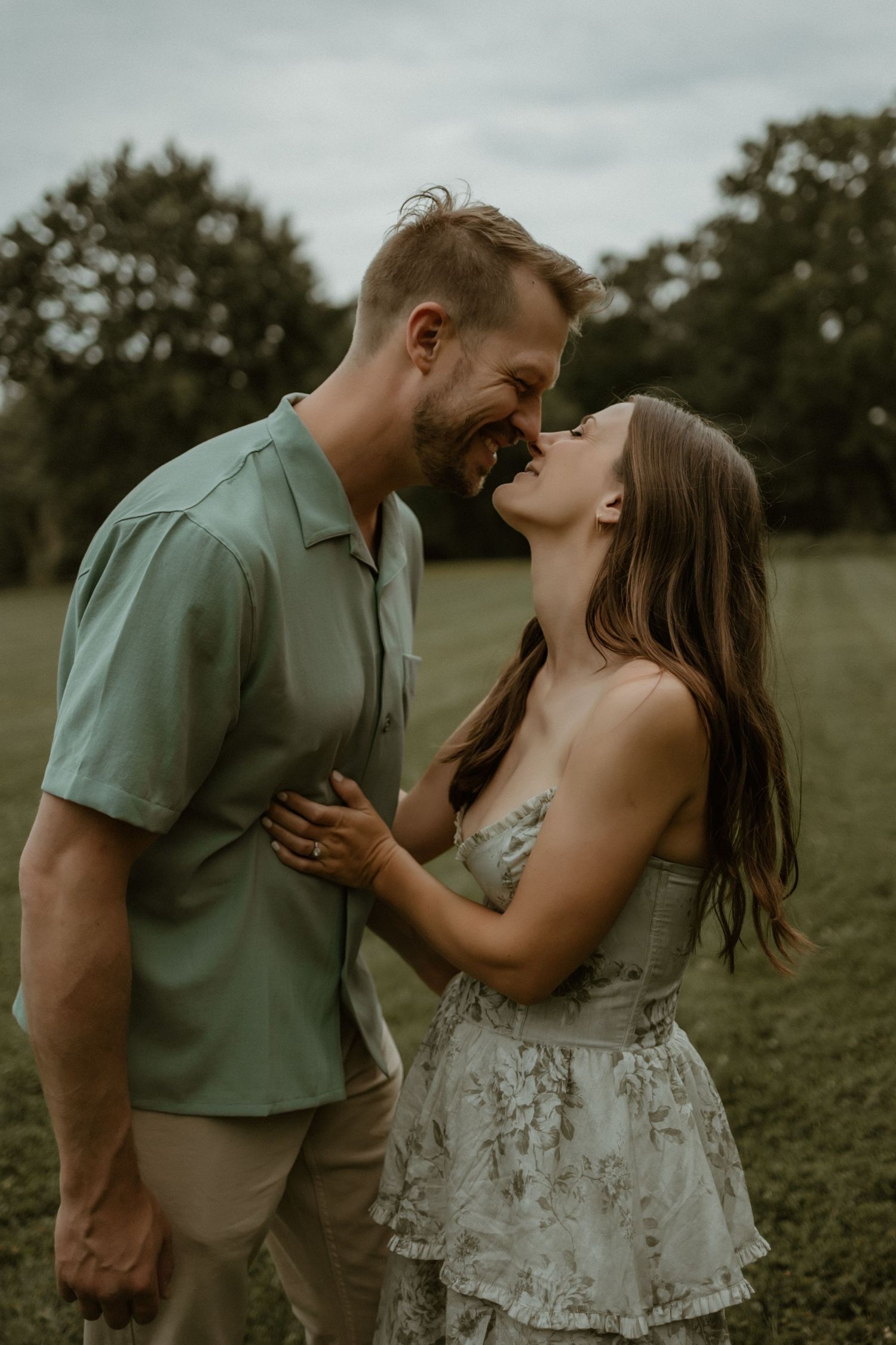 A couple shares a romantic moment laughing together in a grassy field at sunset with moody dark lighting.