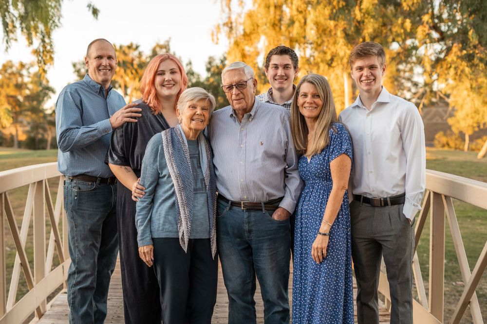 Multi-generation family group smiling together on wooden bridge during golden sunset in autumn.