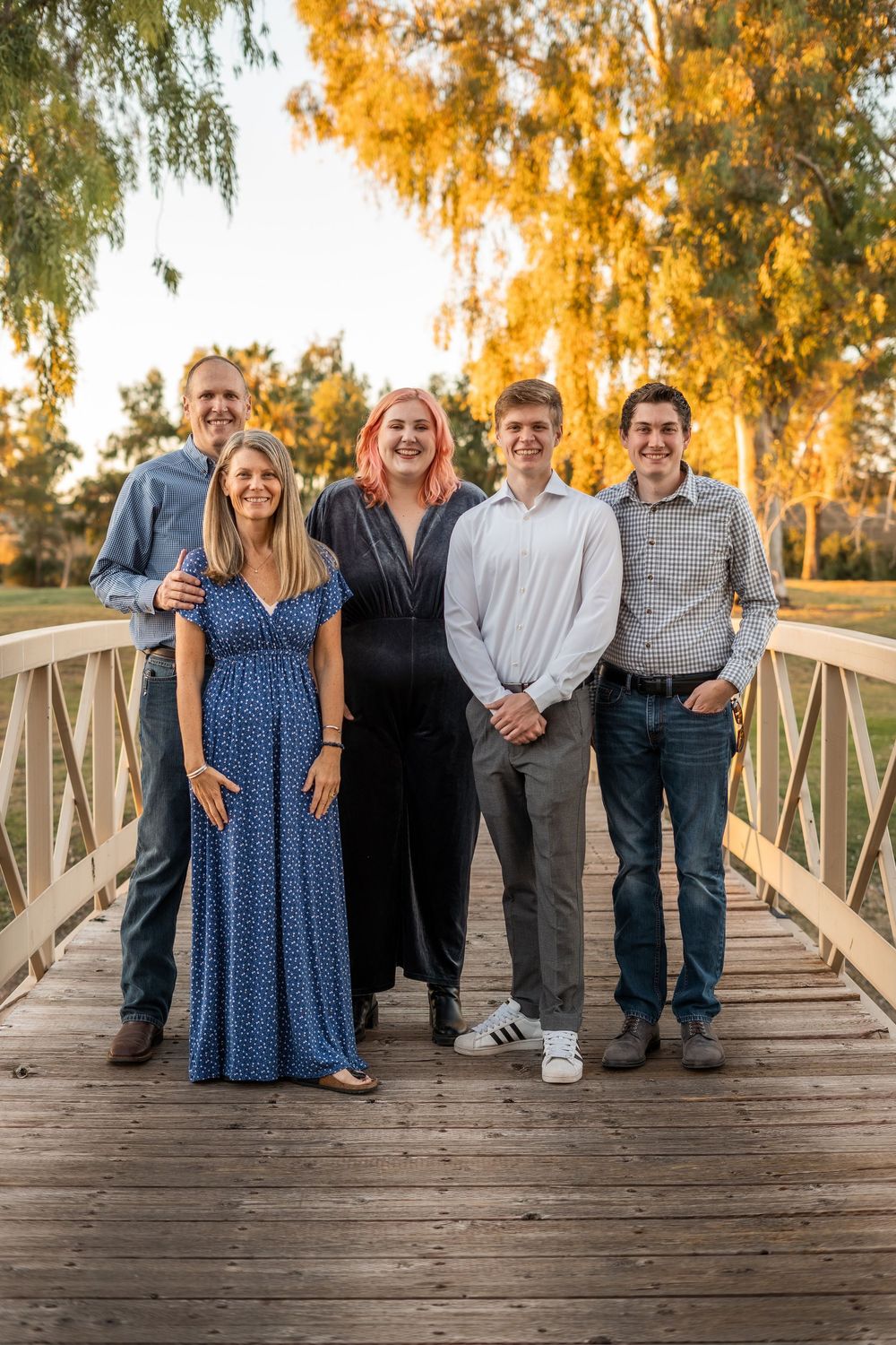 A group of people stand together on a wooden bridge during golden hour with autumn trees in the background.