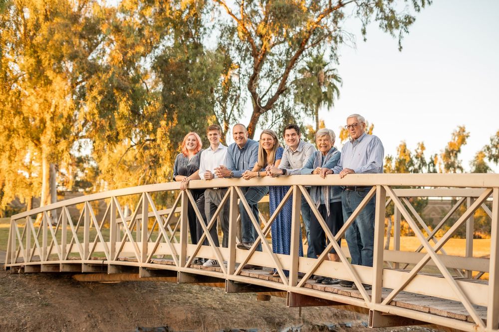 A group of people stand on a wooden footbridge at sunset with golden light filtering through trees.