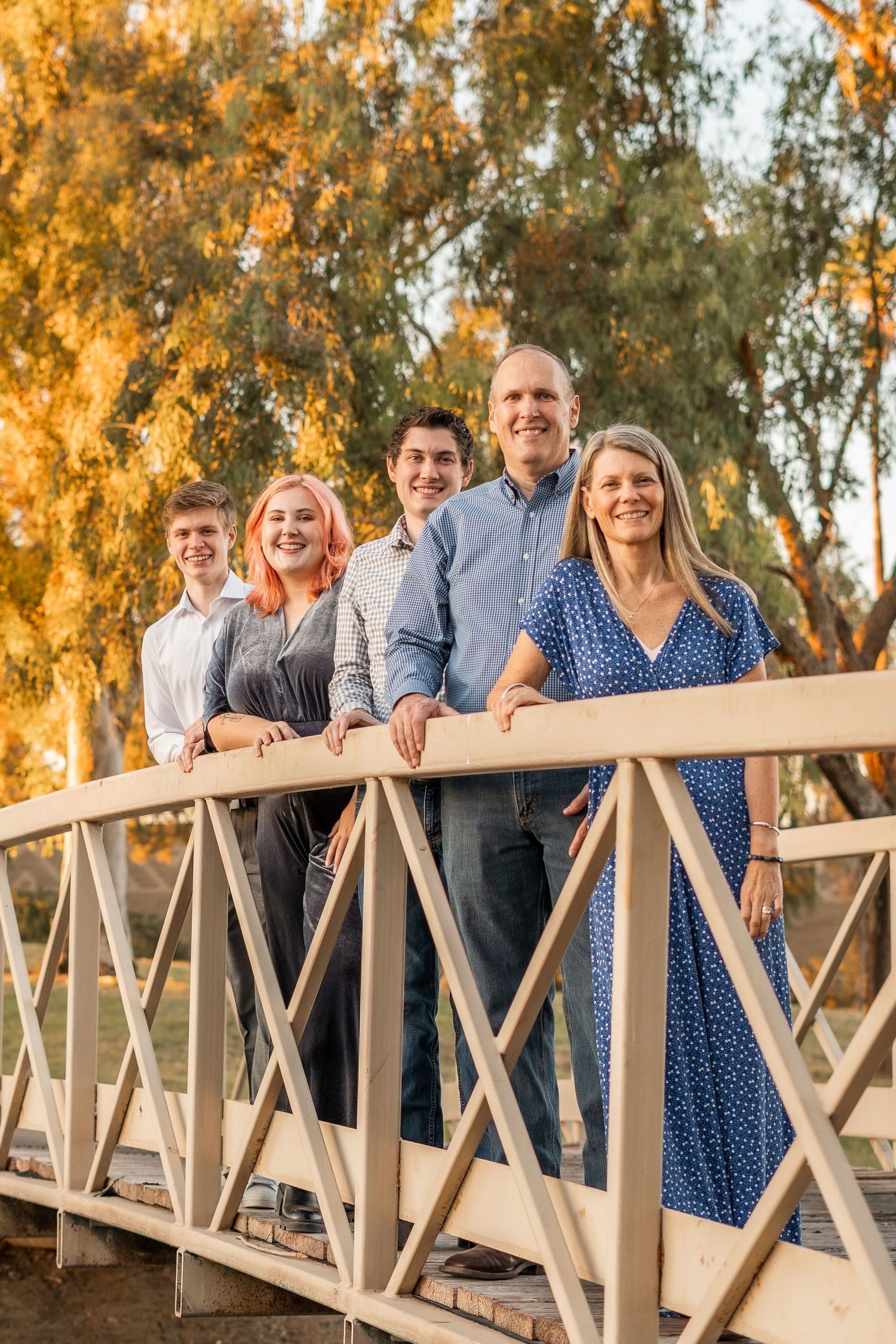 Multi-generational family stands together smiling on a wooden bridge during golden sunset hour.