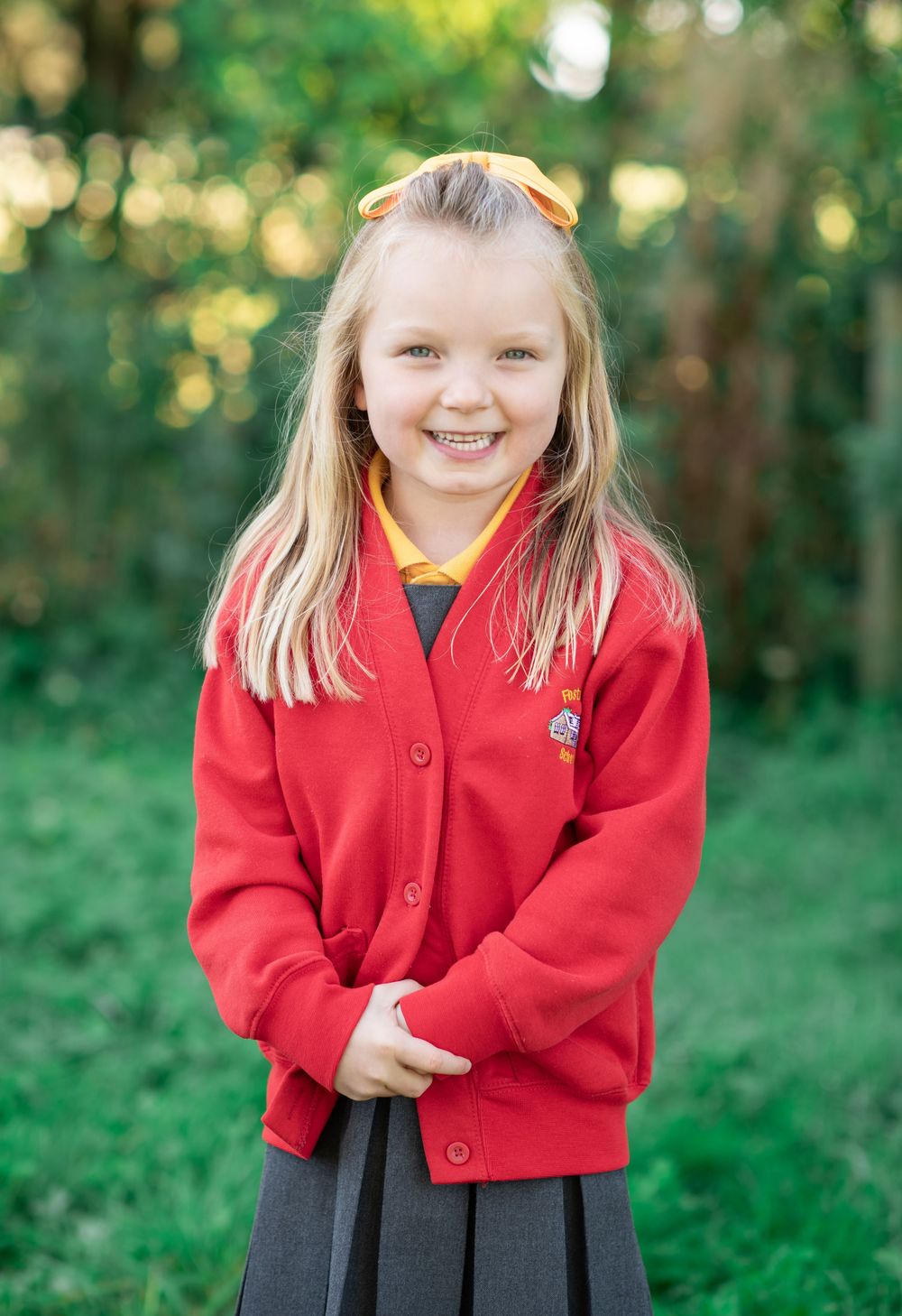 Little girl in her school uniform stands outdoors against blurred green foliage background.
