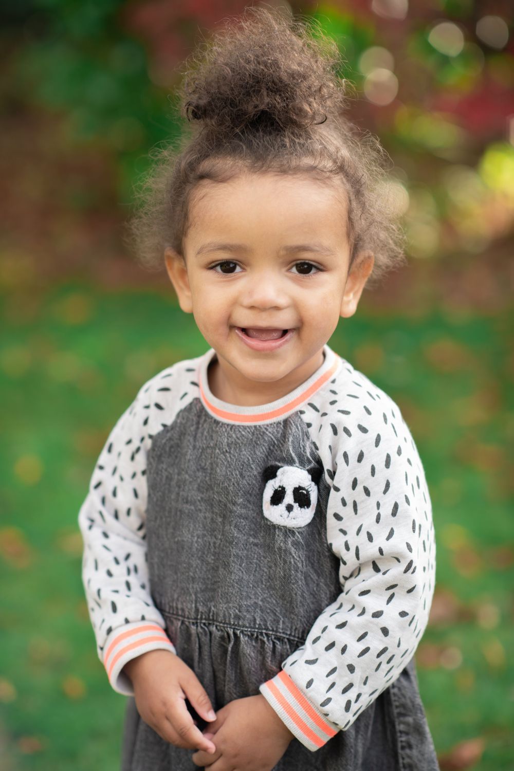 A toddler wearing a panda-themed gray jumper dress with white patterned long sleeves stands outdoors in a garden setting.