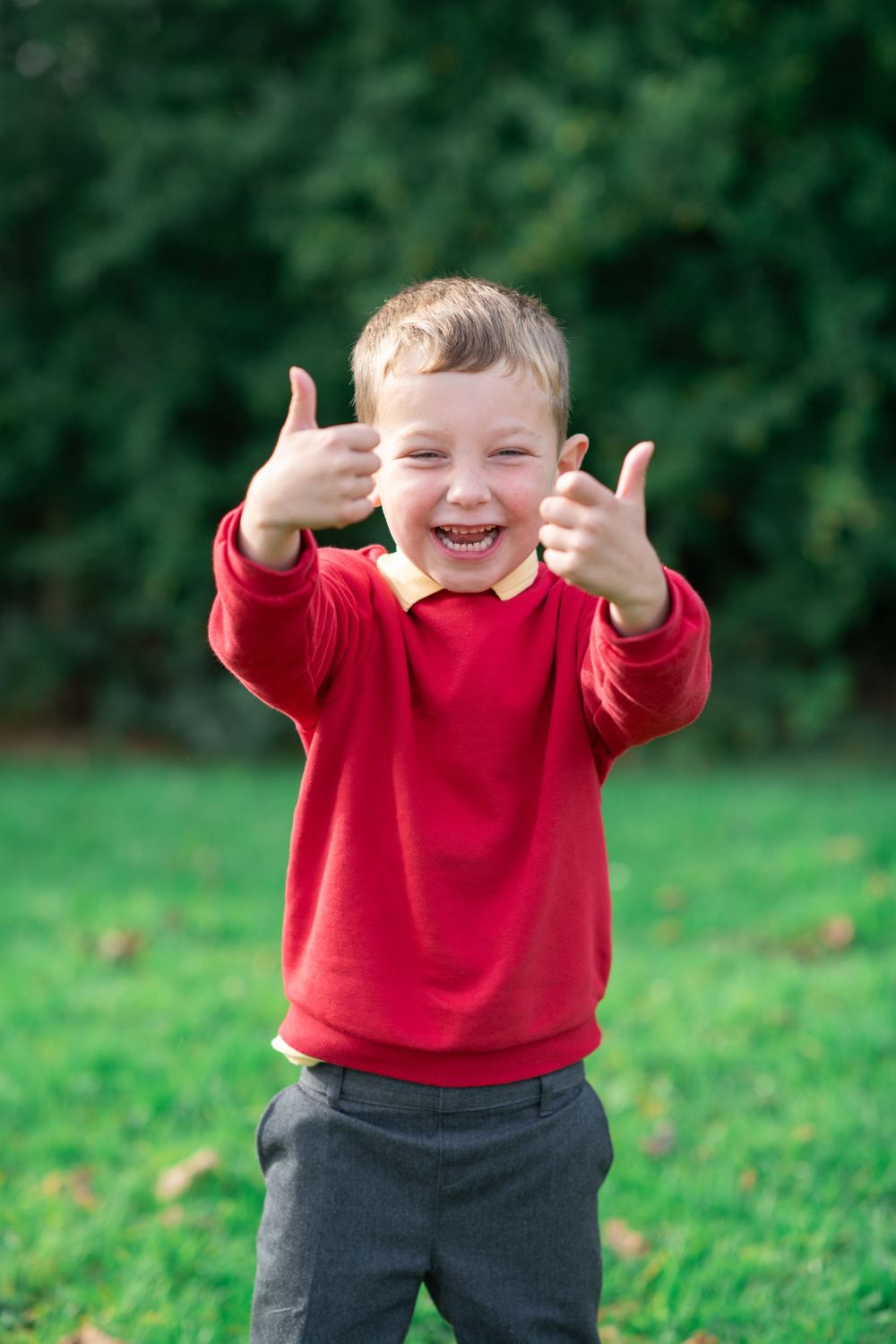 A young child in a red sweater and gray pants stands on grass showing excitement with raised arms and a joyful expression.