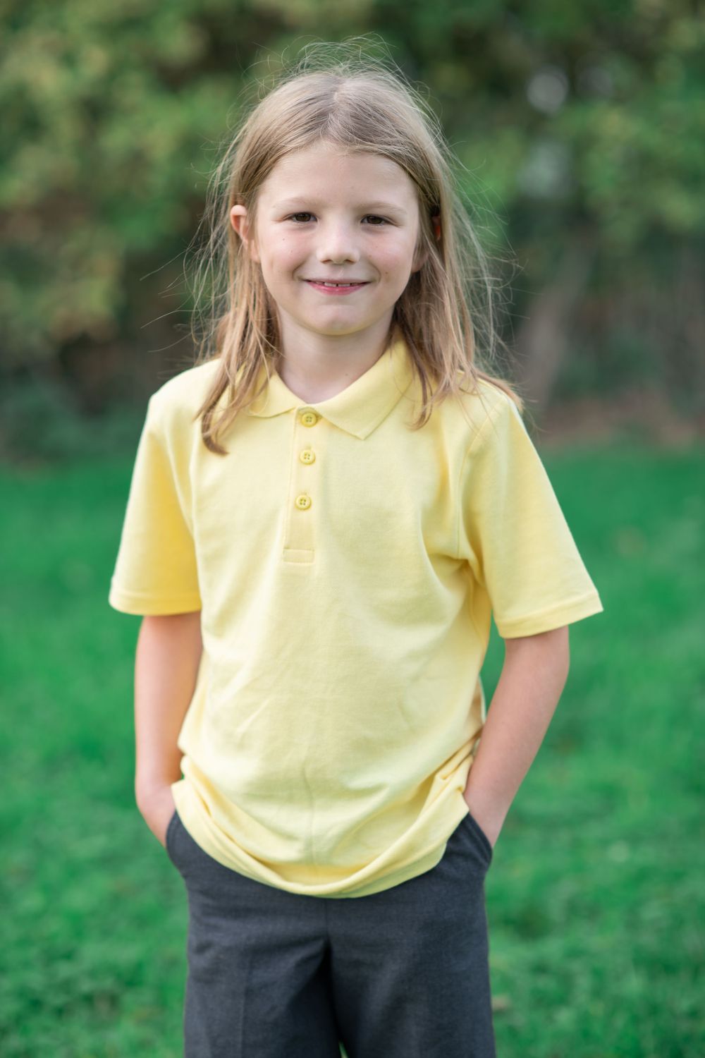 A young student in a yellow polo shirt and dark pants stands on green grass outdoors.