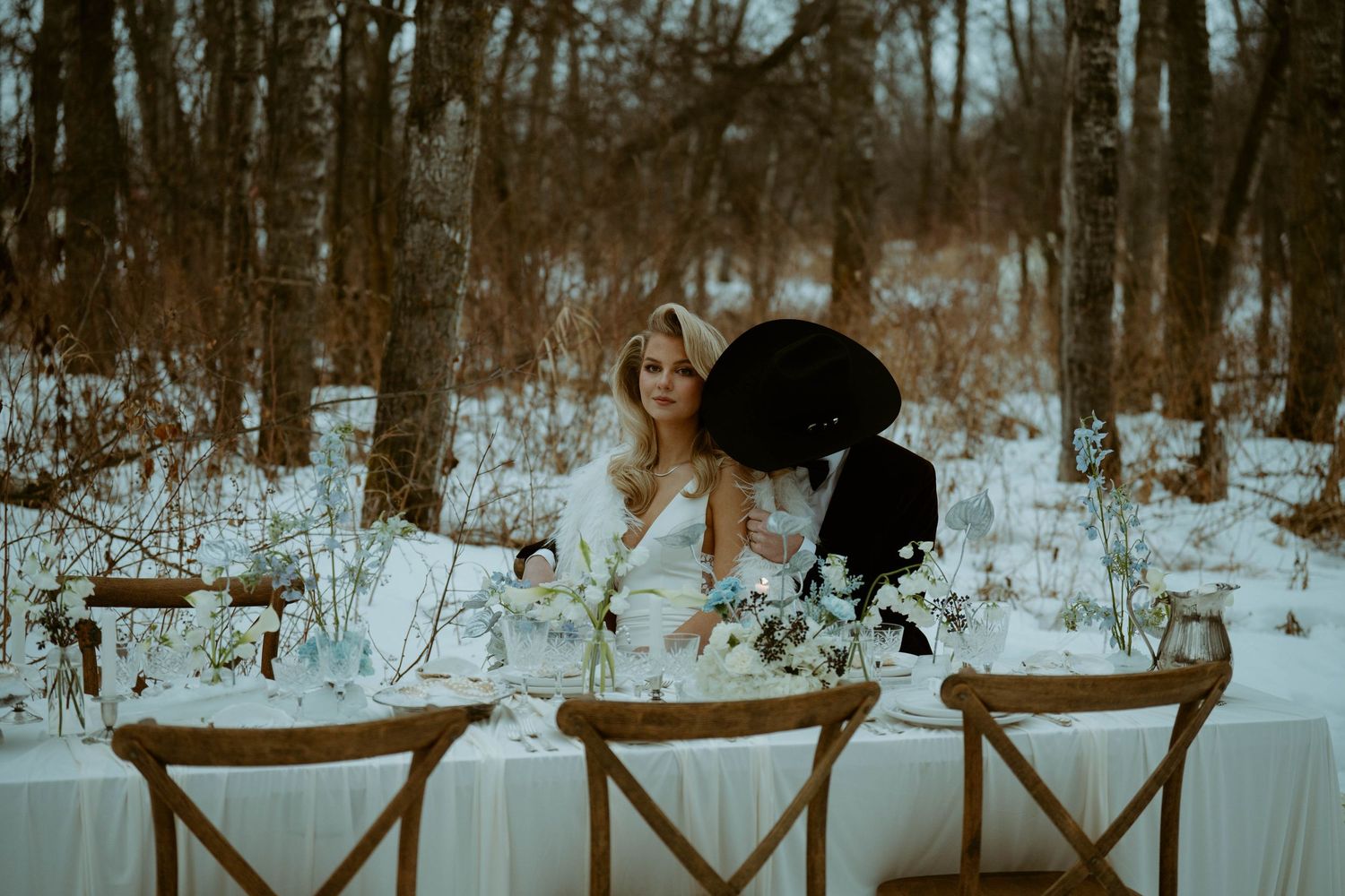 Couple sits at an elegant outdoor winter wedding table surrounded by snow-covered trees and white floral centerpieces.