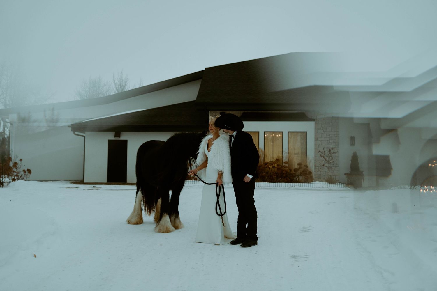Bride and groom sharing a kiss in snow beside a dark horse outside a modern building in winter.