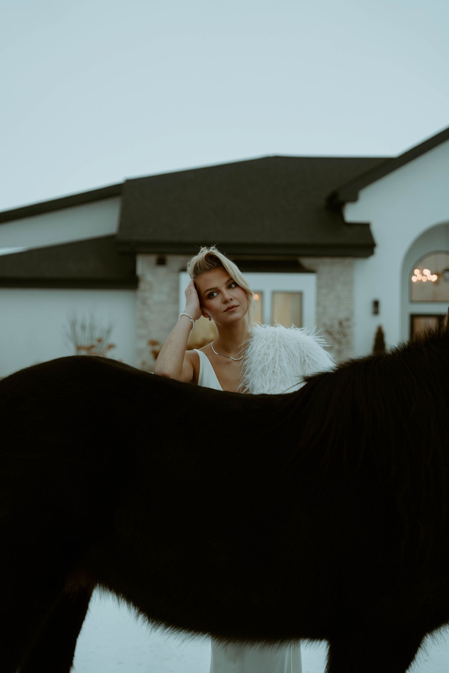 Elegant bride in white dress with feather wrap stands beside a dark horse outside a modern white estate.