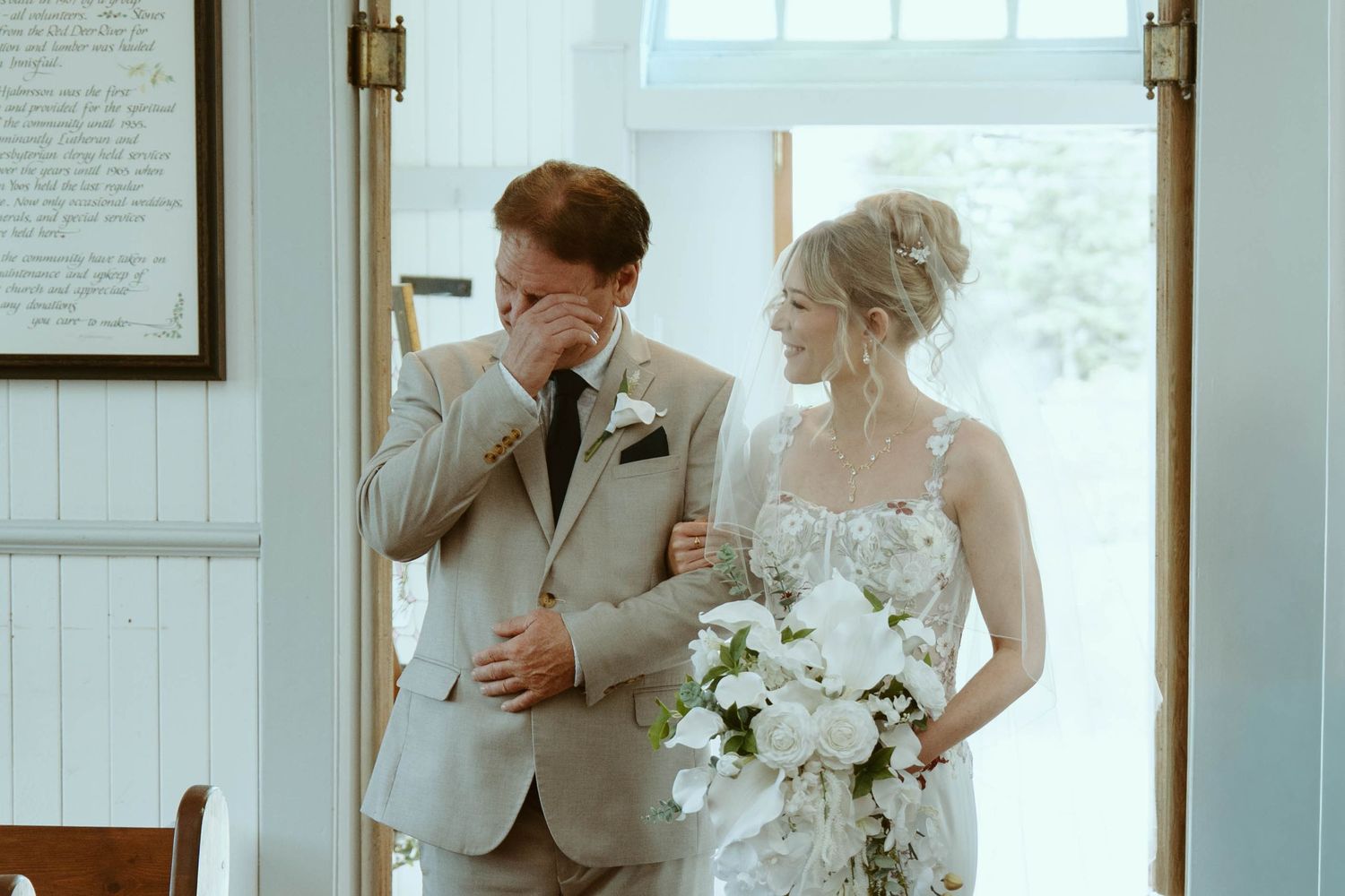 Emotional groom wiping tears beside smiling bride holding white floral bouquet at wedding ceremony entrance.
