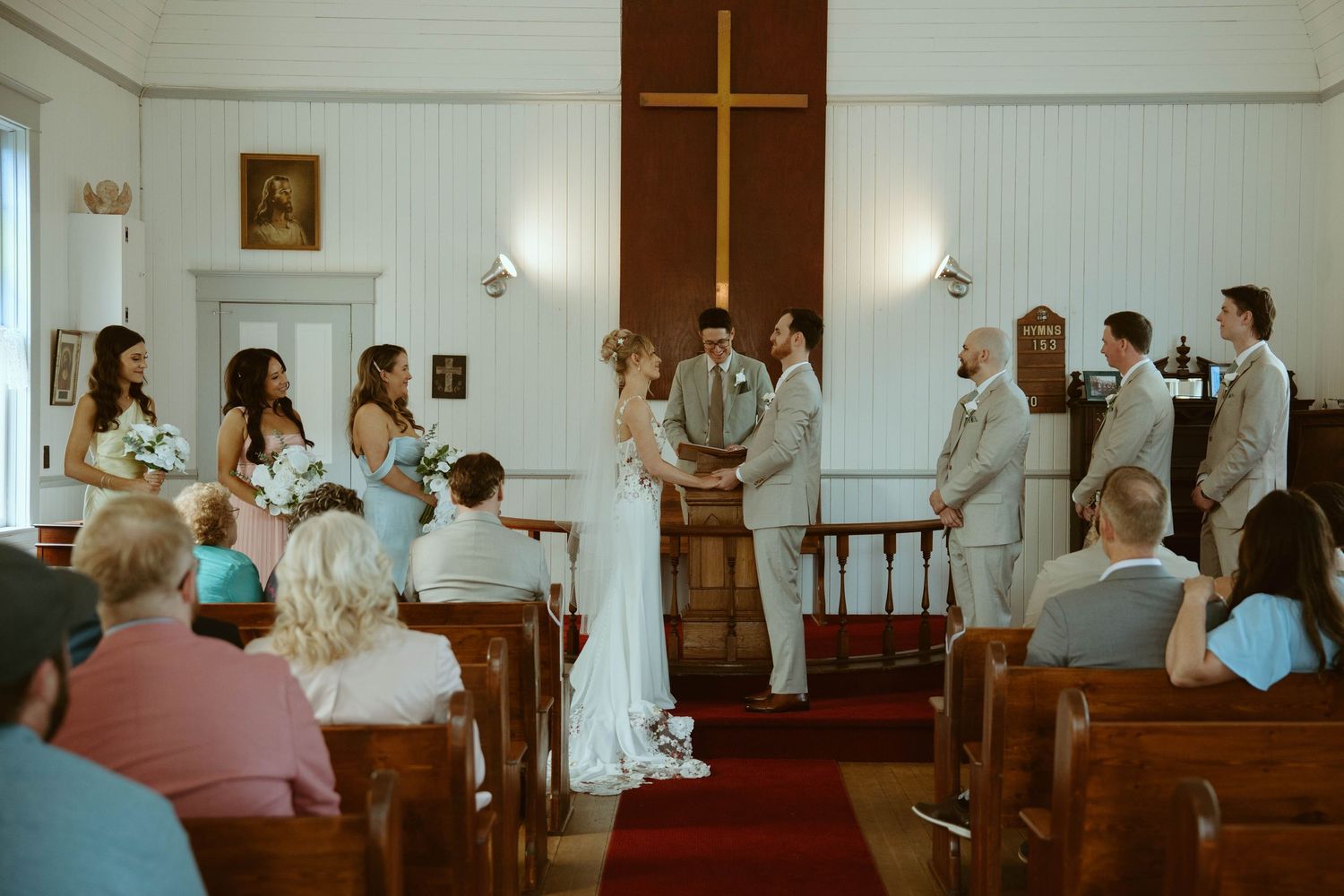 Bride and groom exchange vows at a small white church altar with wooden cross, surrounded by wedding party and guests.