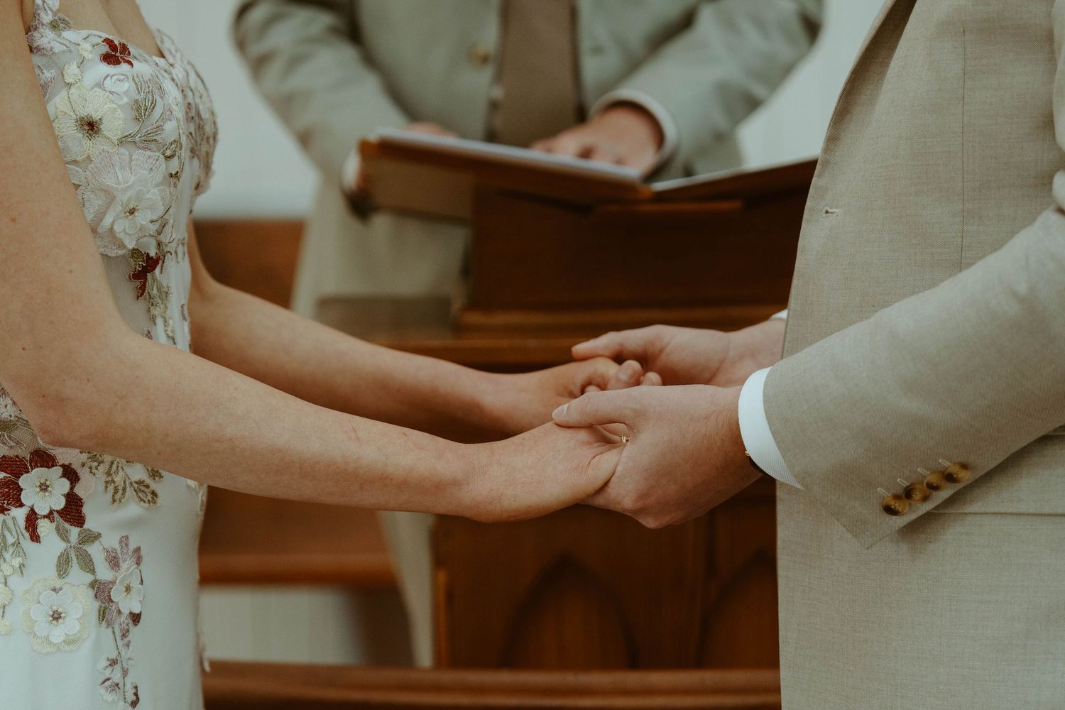 Couple holding hands at wedding altar with officiant and wooden pews in background.