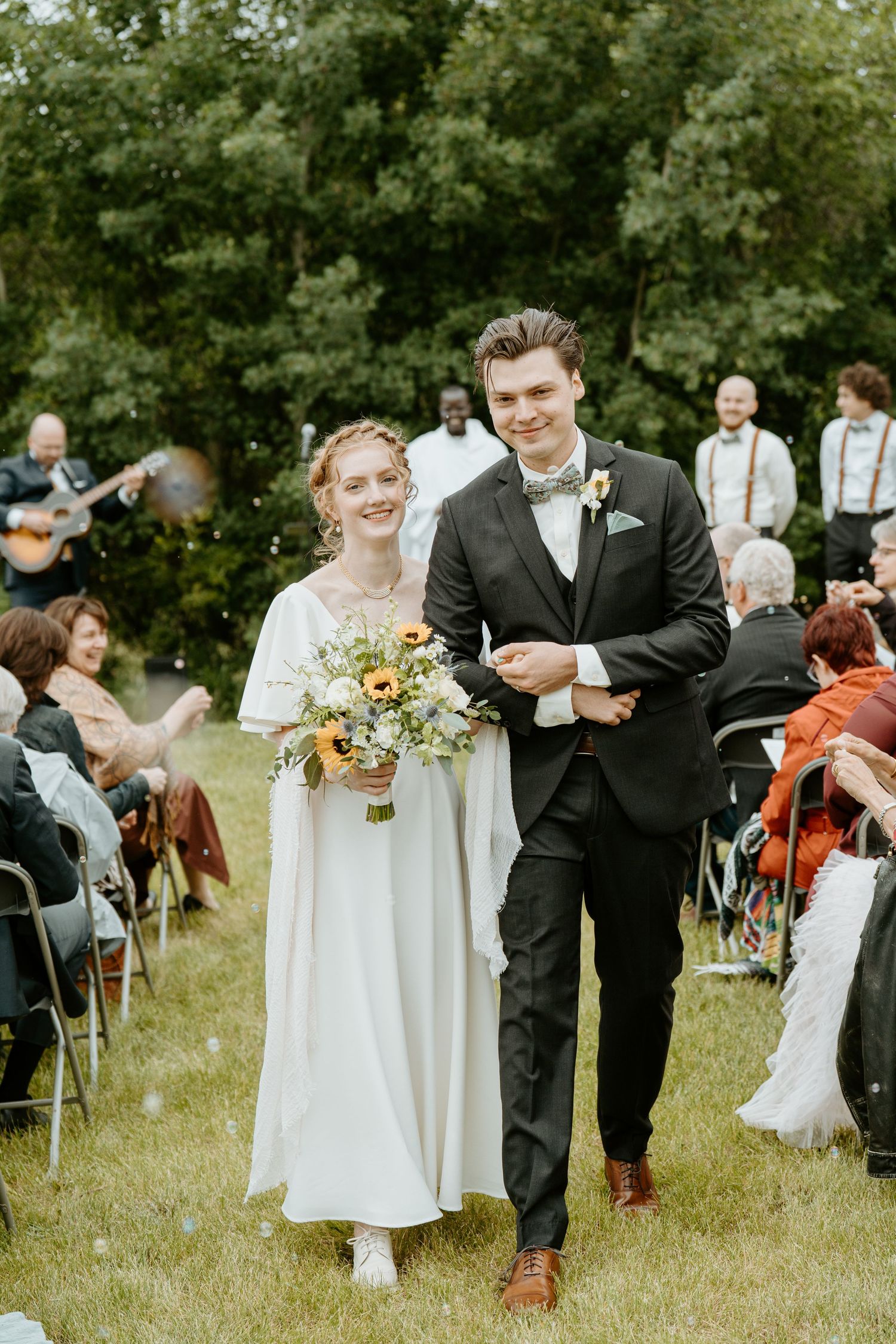 A joyful outdoor wedding ceremony in a garden setting with guests seated on chairs and musicians playing in the background.