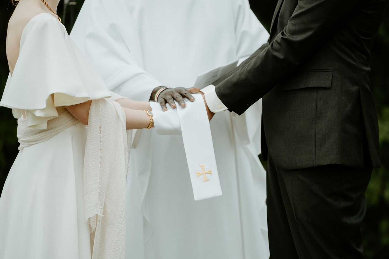 Close-up shot of hands holding during wedding ceremony with white dress and dark suit details.