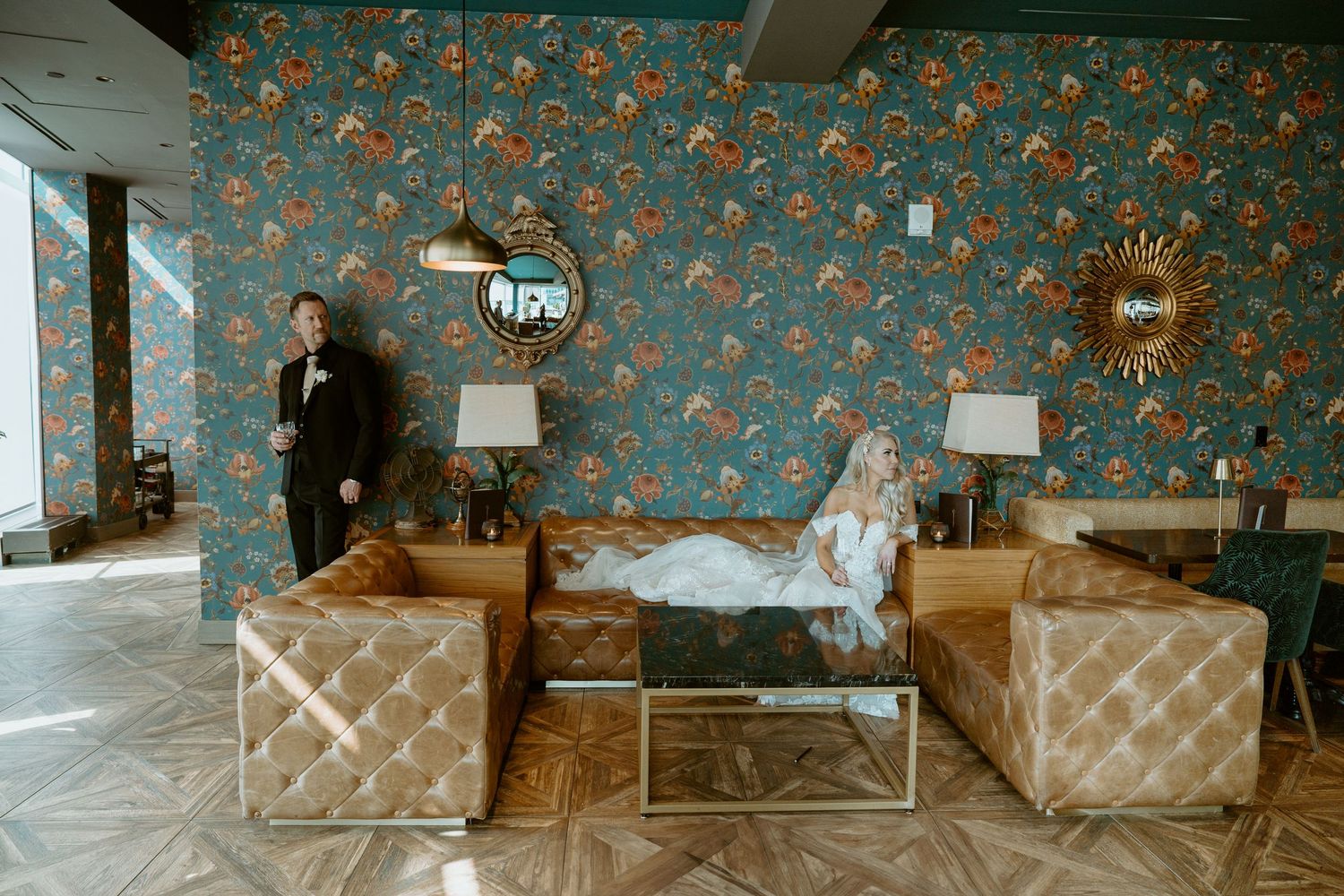 Vintage living room with blue floral wallpaper features brown leather tufted sofa and coordinating armchair.